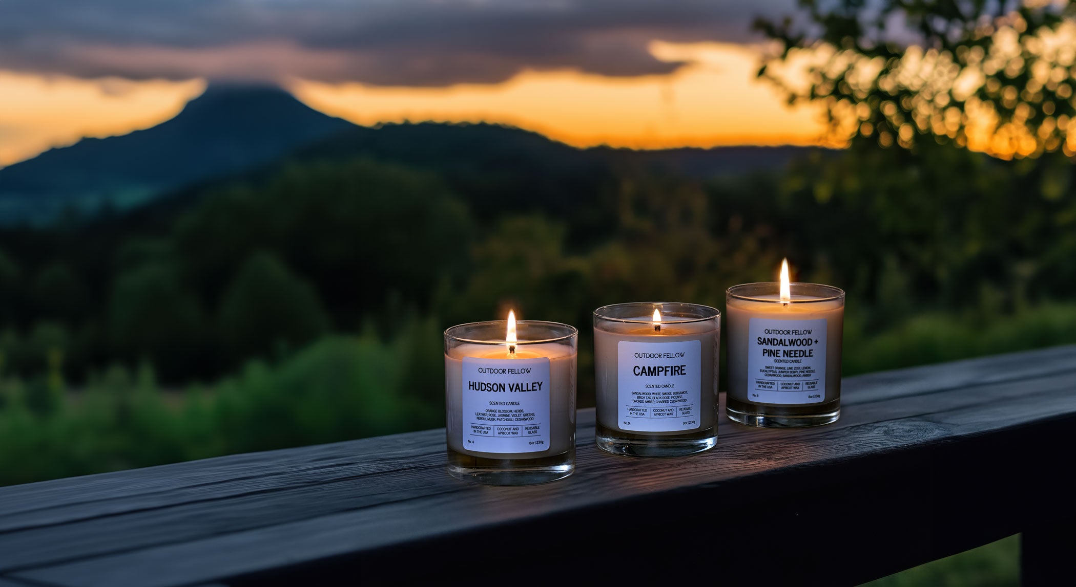 Three lit Outdoor Fellow candles on a railing at sunset with mountains in the background.