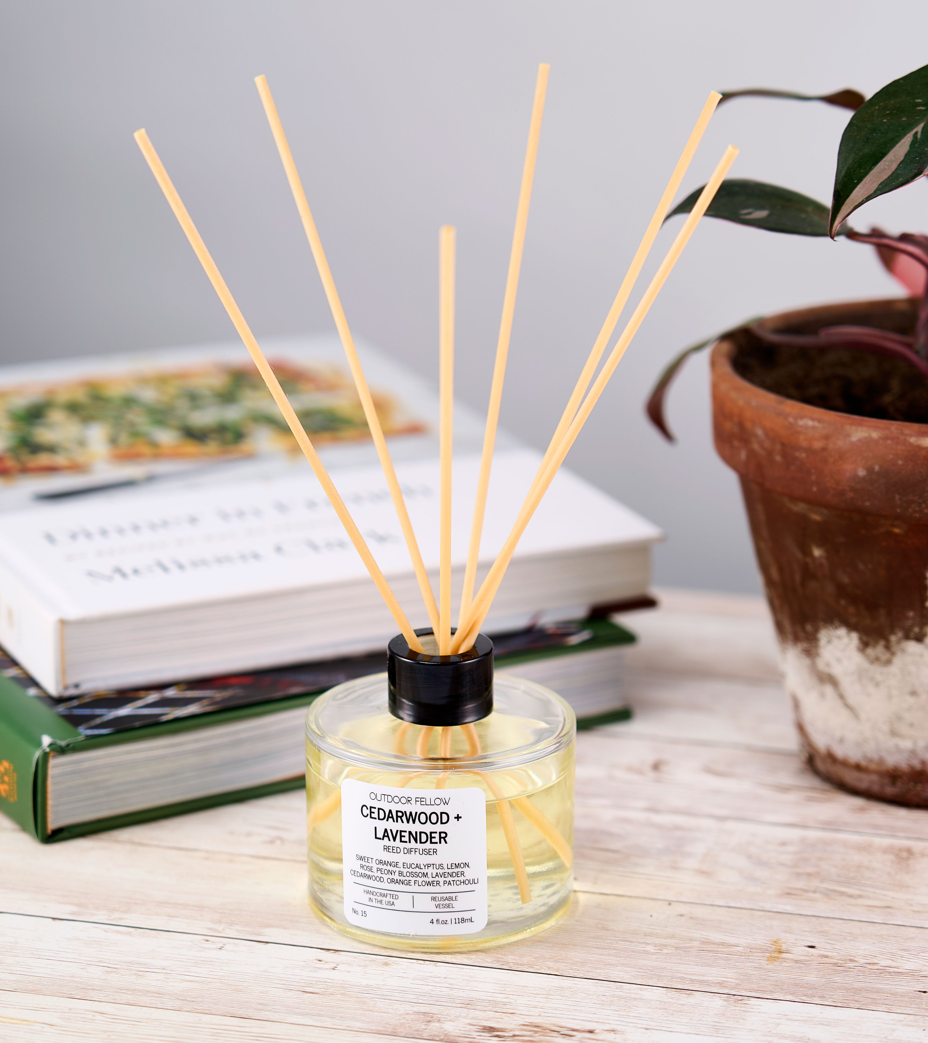 Outdoor Fellow Cedarwood + Lavender reed diffuser on a rustic wood table with stacked cookbooks and a potted plant in the background.