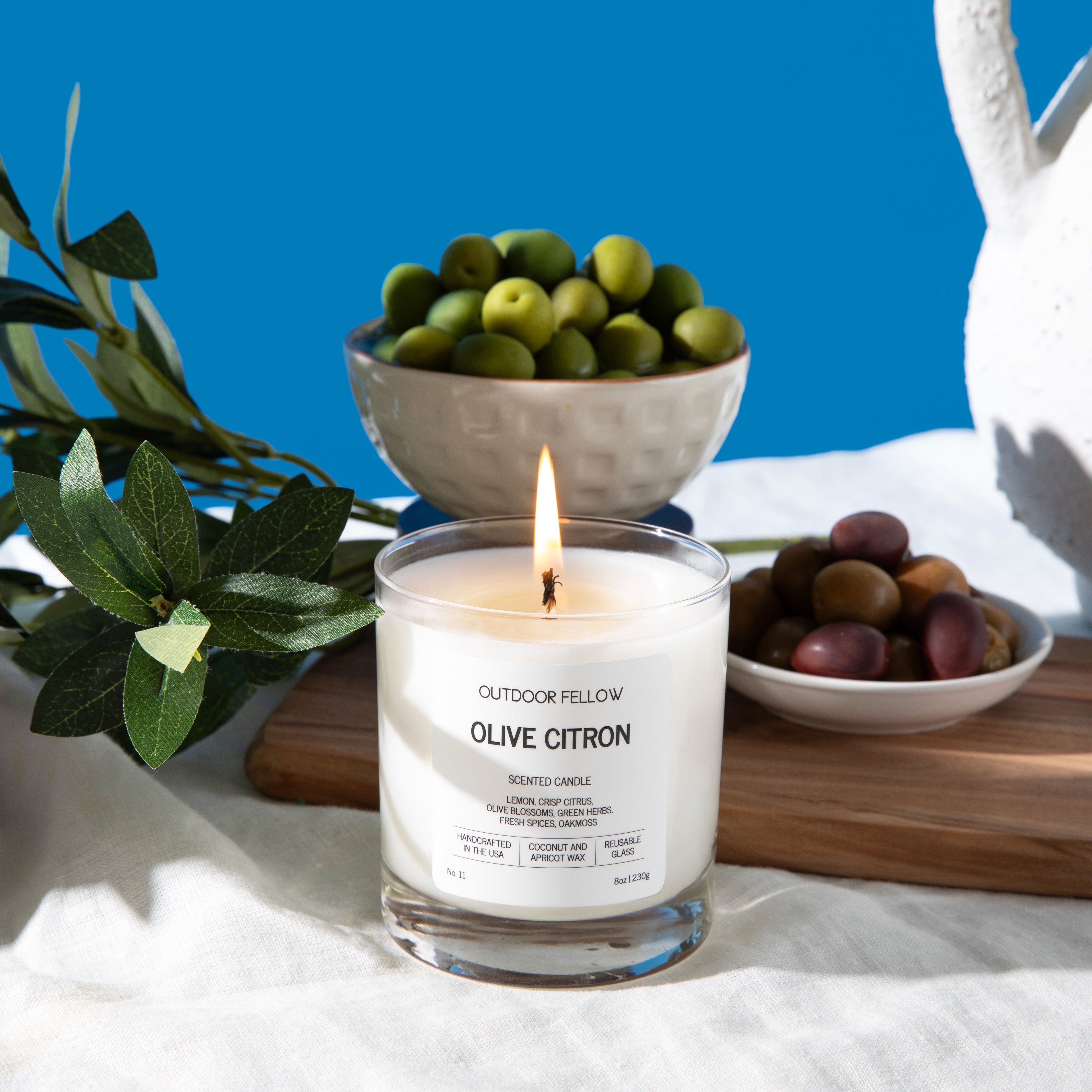 A lit Outdoor Fellow Olive Citron scented candle sits on a white linen cloth surrounded by Mediterranean-inspired elements. To the left, a bowl of lemons and sprigs of olive branches with dark olives rest on a wooden surface. Behind the candle are ceramic bowls filled with green and multicolored olives, and to the right is a white, textured ceramic pitcher. The backdrop is a vibrant blue, creating a bright and fresh atmosphere.