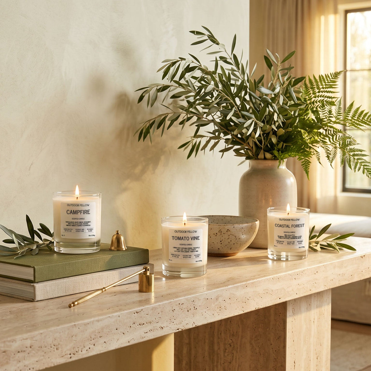 Three lit candles on a marble table beside books, olive branches, and a leafy vase.