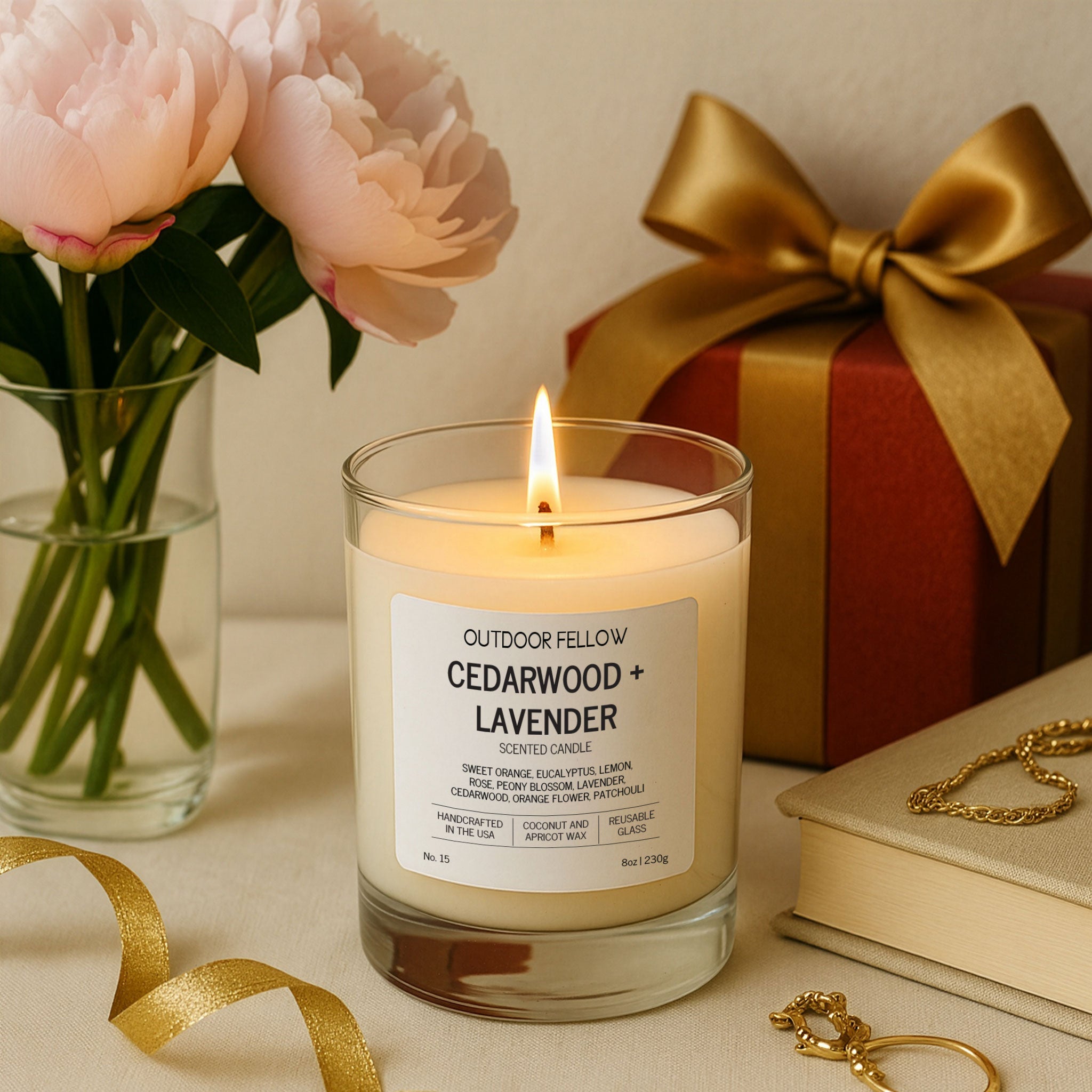 A lit Outdoor Fellow Cedarwood + Lavender candle sits on a cream-colored surface beside a red gift box tied with a gold ribbon, a vase of soft pink peonies, and delicate gold jewelry.