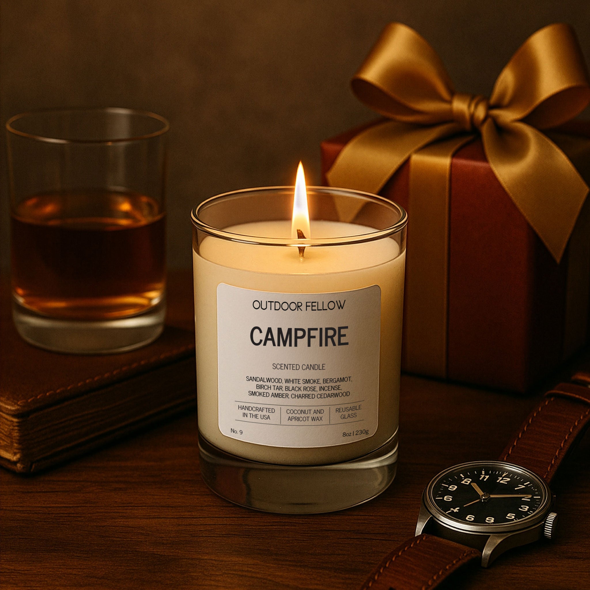 A lit Outdoor Fellow Campfire candle sits on a dark wooden surface beside a glass of whiskey, a brown leather watch, and a gift box wrapped with a gold ribbon.