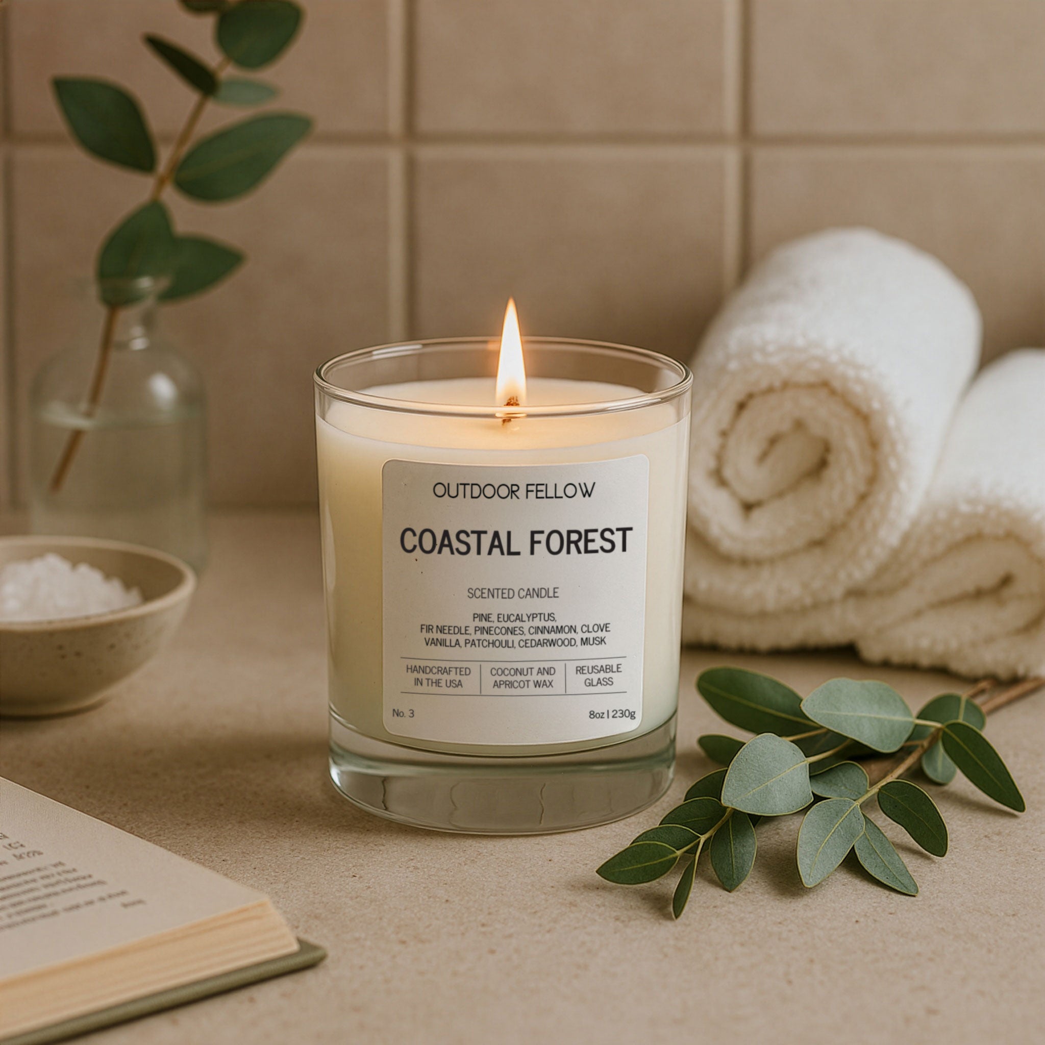 A lit Outdoor Fellow Coastal Forest candle sits on a spa-like surface beside rolled white towels, eucalyptus leaves, and a small bowl of bath salts, creating a calm and soothing atmosphere.