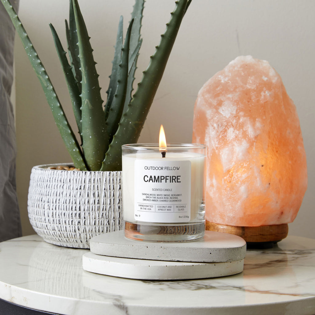 Candle labeled 'Campfire' with a potted plant and a Himalayan salt lamp on a marble surface.