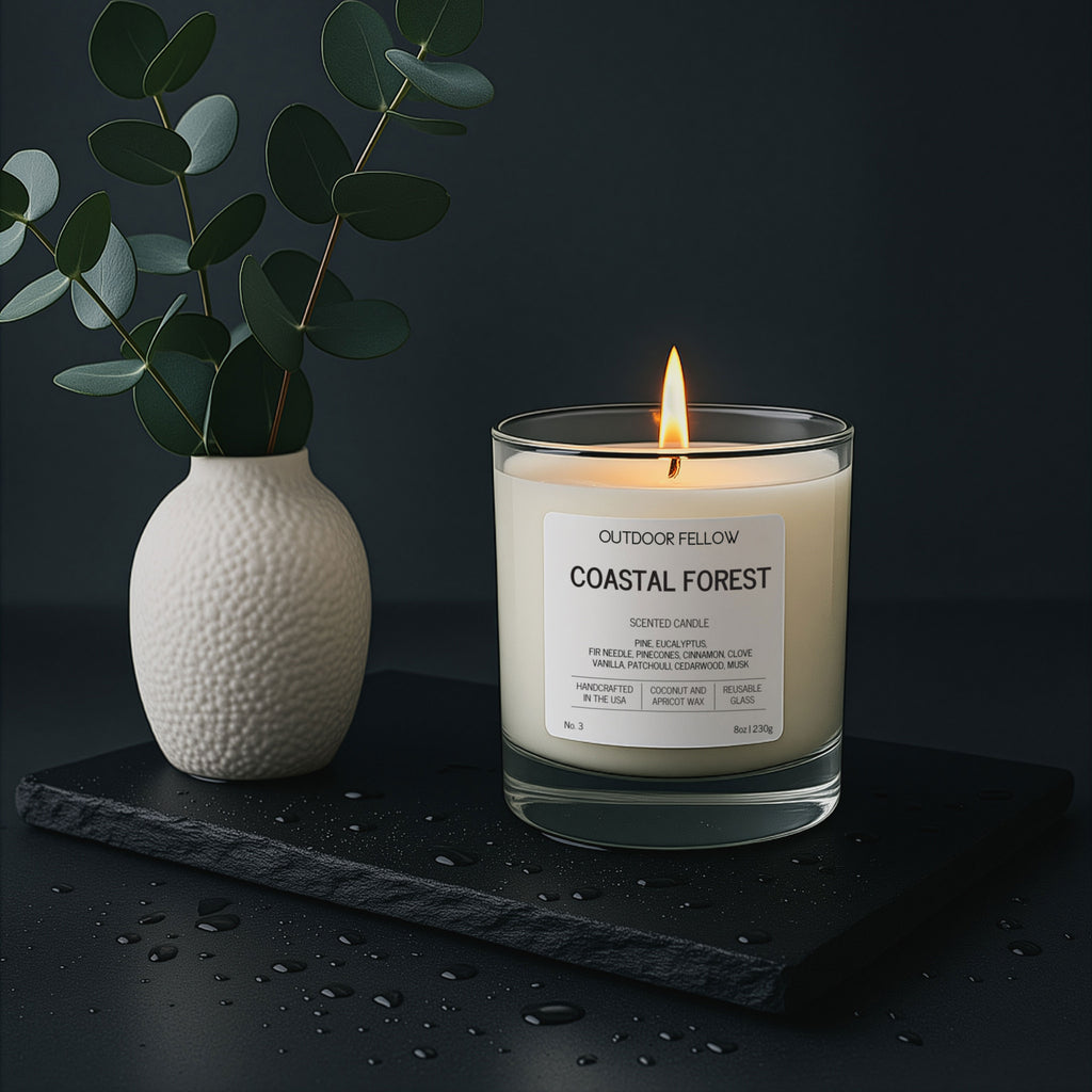 Candle labeled 'Coastal Forest' by Outdoor Fellow on a dark surface with a vase of greenery.