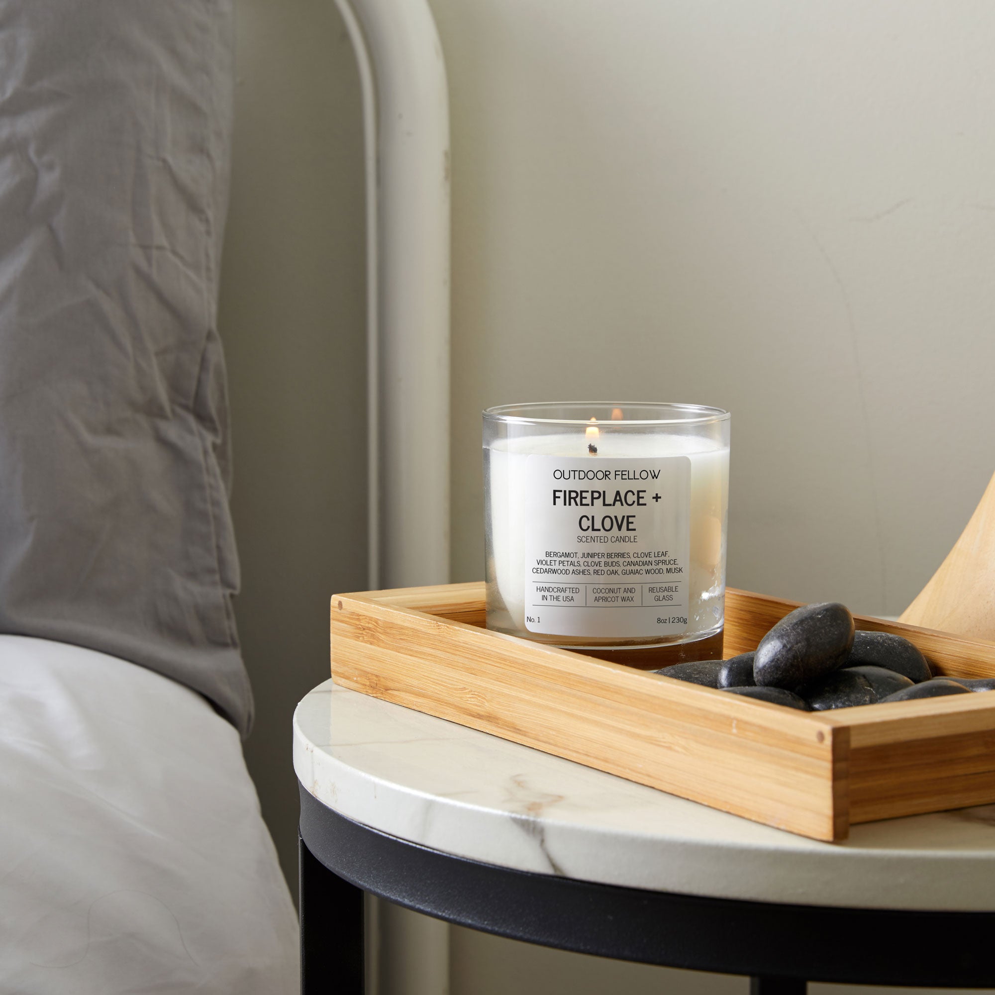 Candle labeled 'Fireplace + Clove' on a marble nightstand with a wooden tray and rocks.