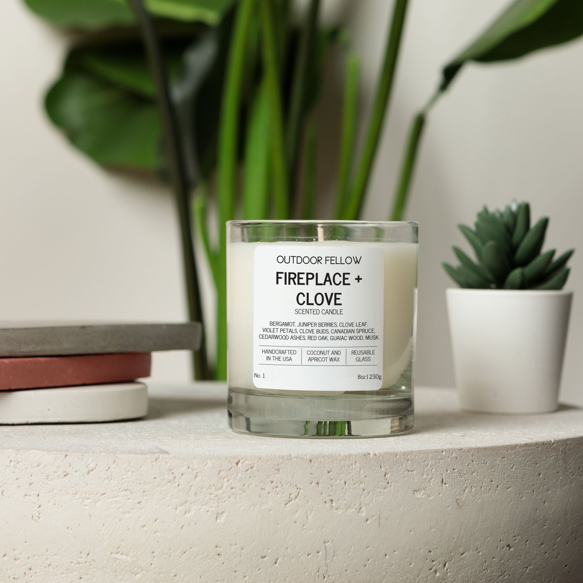 Candle labeled 'Fireplace + Clove' by 'Outdoor Fellow' on a shelf with plants in the background.