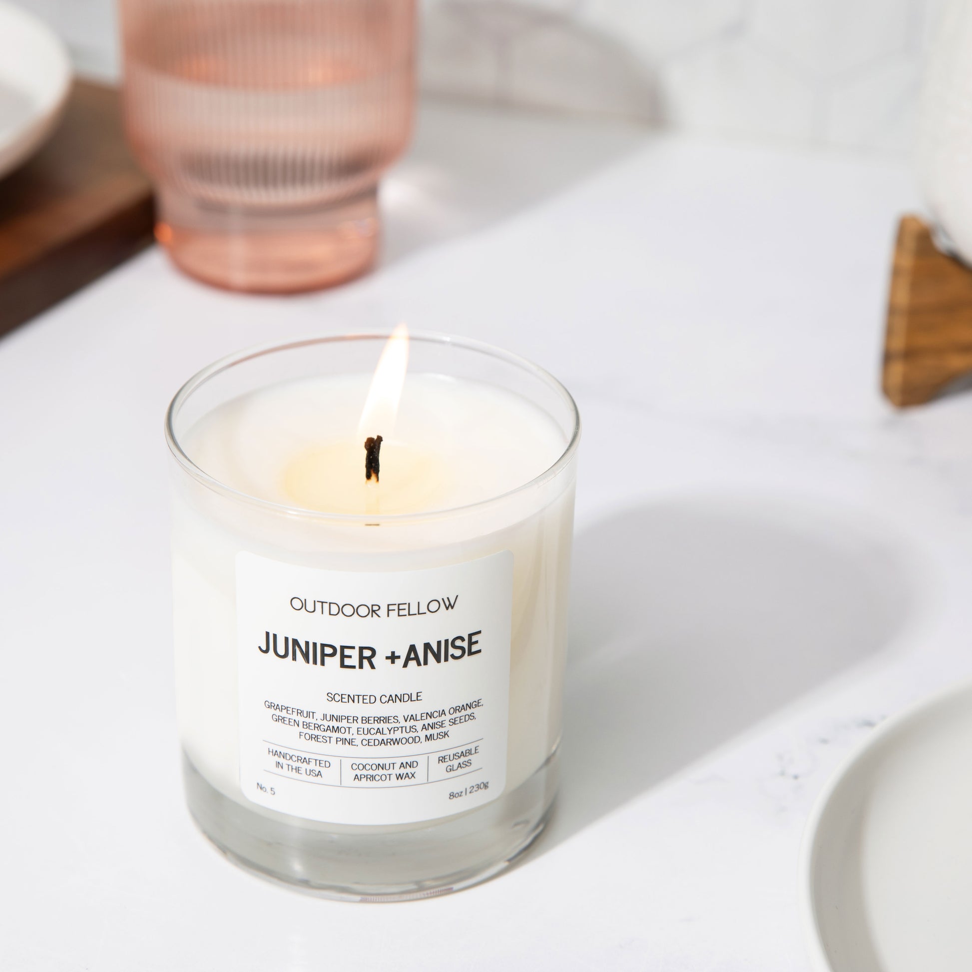 Juniper and Anise scented candle on a white surface with a pink water glass in the background. 