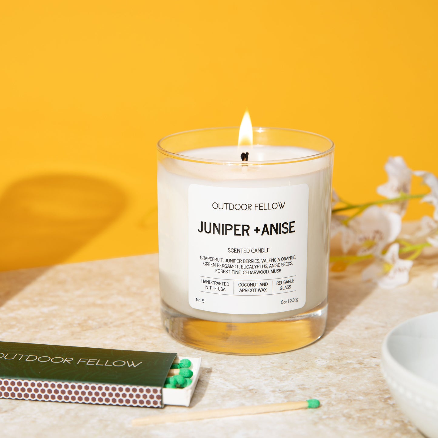 Juniper and Anise scented candle in front of a yellow background with a box of matches