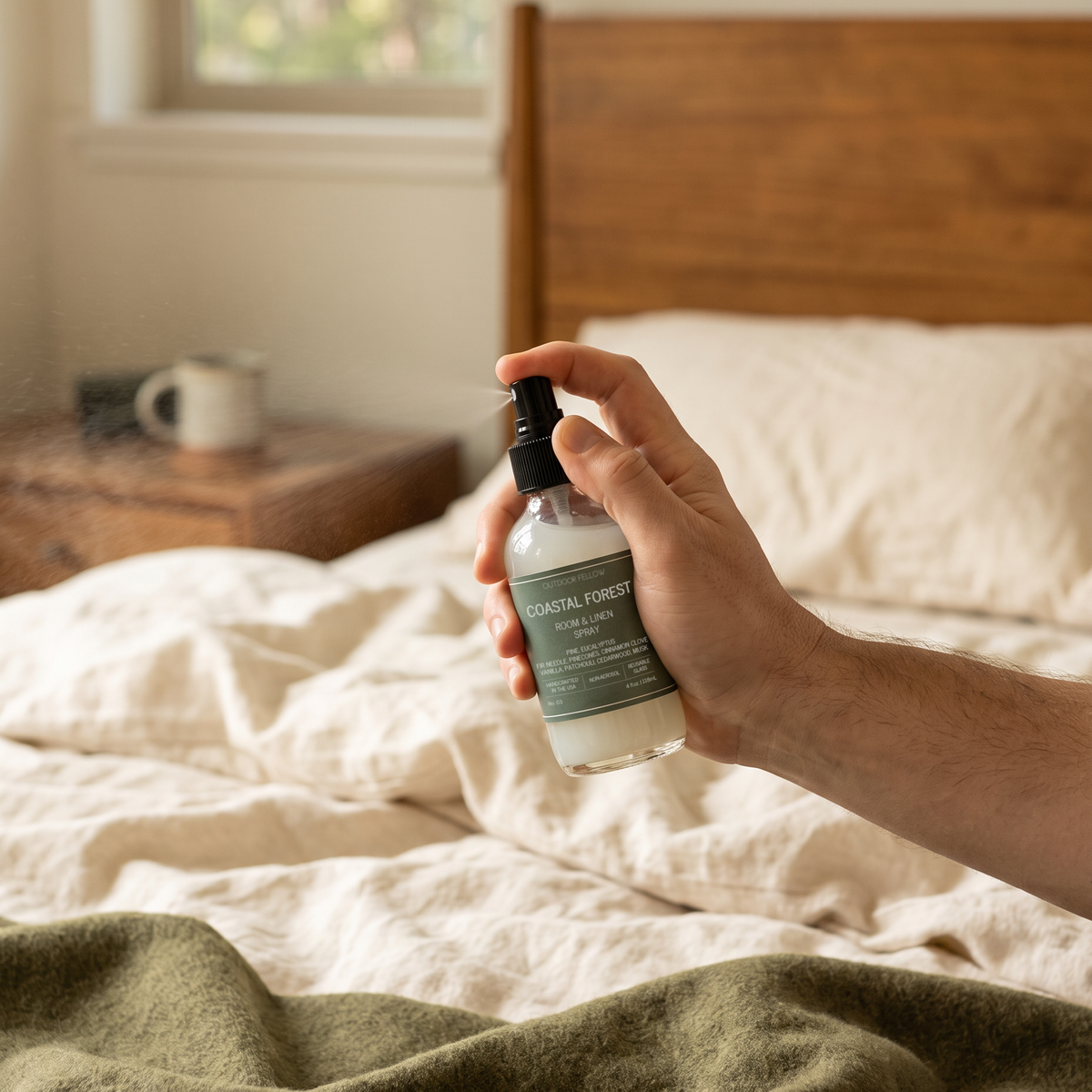 Hand spraying Outdoor Fellow Coastal Forest Room & Linen Spray over a bed with natural linen bedding and a green throw.