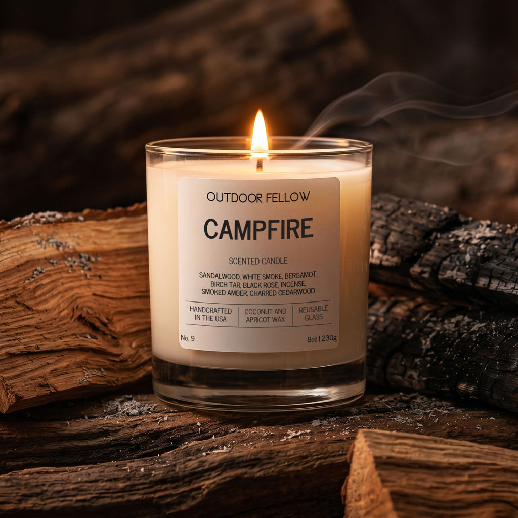 Outdoor Fellow Campfire candle surrounded by charred firewood and ash, with a warm flame and soft smoke rising.