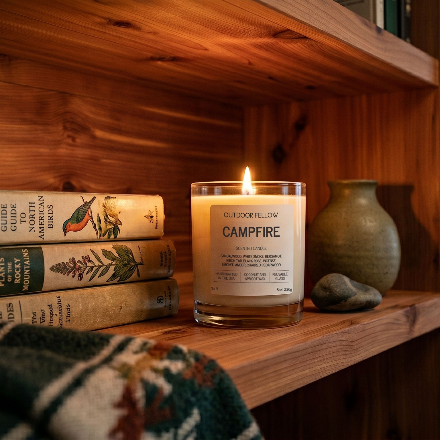 Outdoor Fellow Campfire candle glowing on a wood shelf beside vintage books, a ceramic vase, and a stone accent.