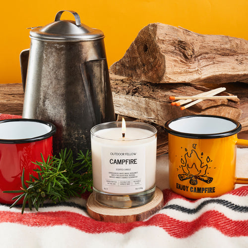 Candle labeled 'Campfire' with camping-themed mugs and a teapot on a wooden surface.
