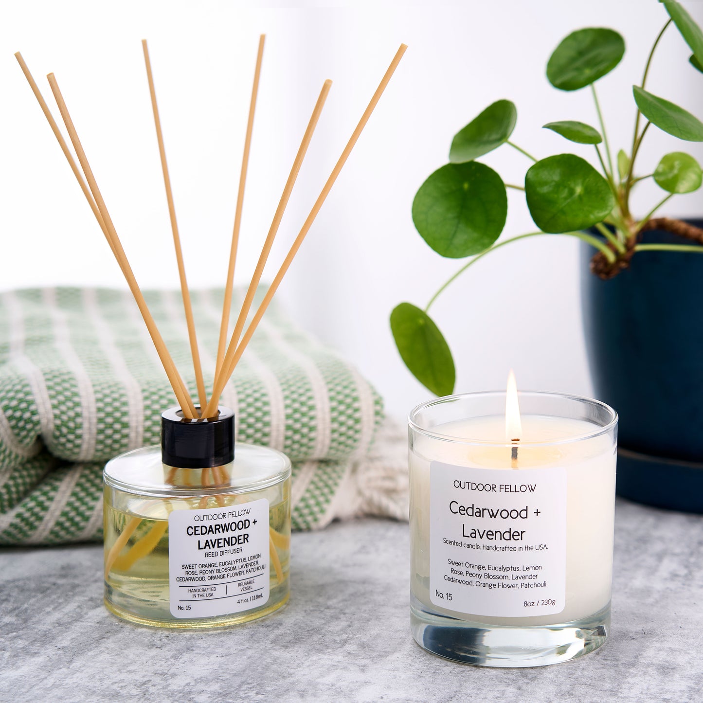 Cedarwood and Lavender reed diffuser and scented candle next to one another with a towel and plant in the background