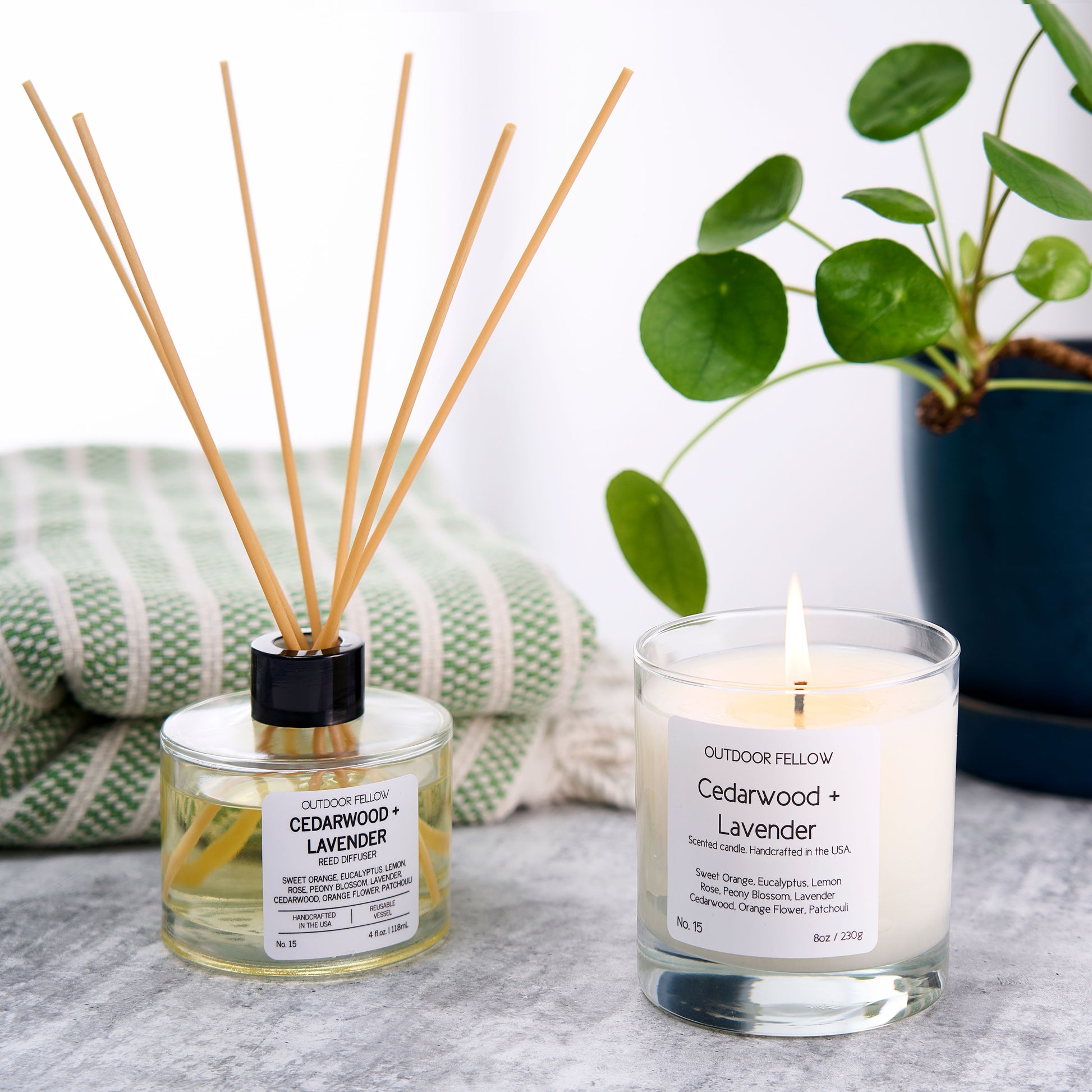 Cedarwood and Lavender reed diffuser and scented candle next to one another with a towel and plant in the background