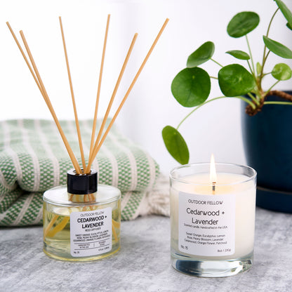 Cedarwood and Lavender reed diffuser and scented candle next to one another with a towel and plant in the background
