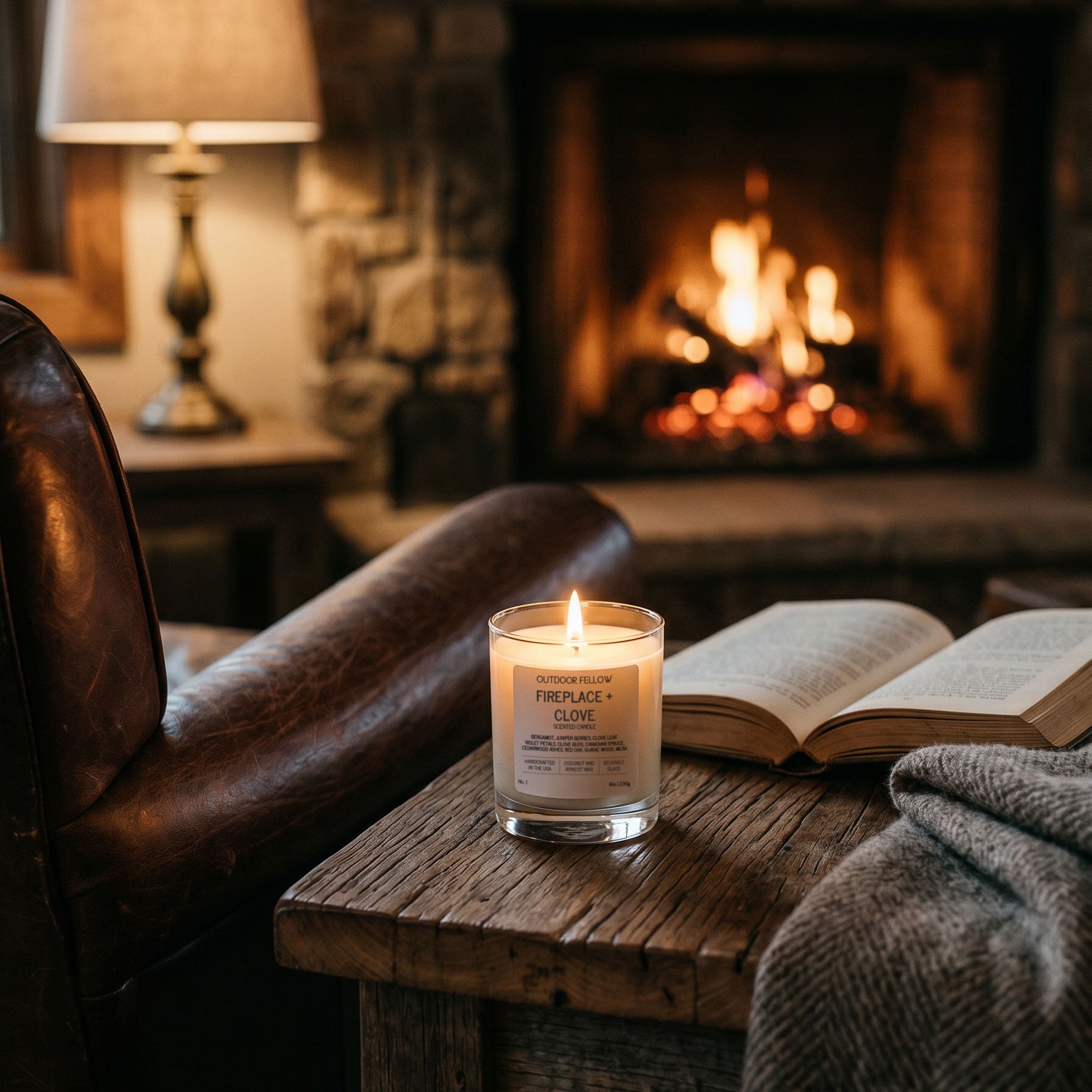 A lit Outdoor Fellow "Fireplace + Clove" scented candle sits on a rustic wooden side table next to an open book. In the soft-focus background, a warm fire crackles in a stone fireplace alongside a leather armchair and a grey knit blanket, evoking a cozy indoor atmosphere.
