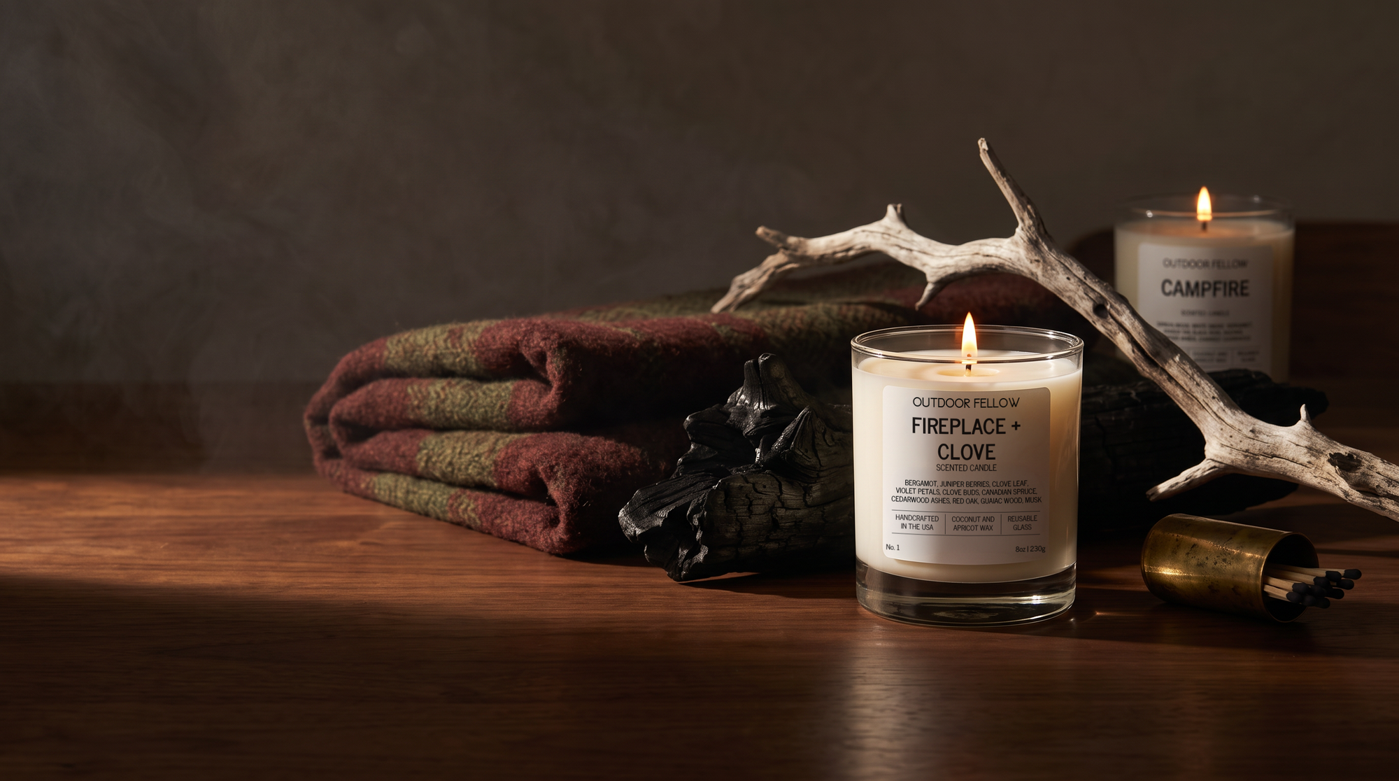 A lit glass jar candle labeled 'Outdoor Fellow Fireplace + Clove' sits on a wooden surface beside a folded patterned blanket, a second lit candle labeled 'Campfire,' a branch, and a small brass container in a moody rustic setting.