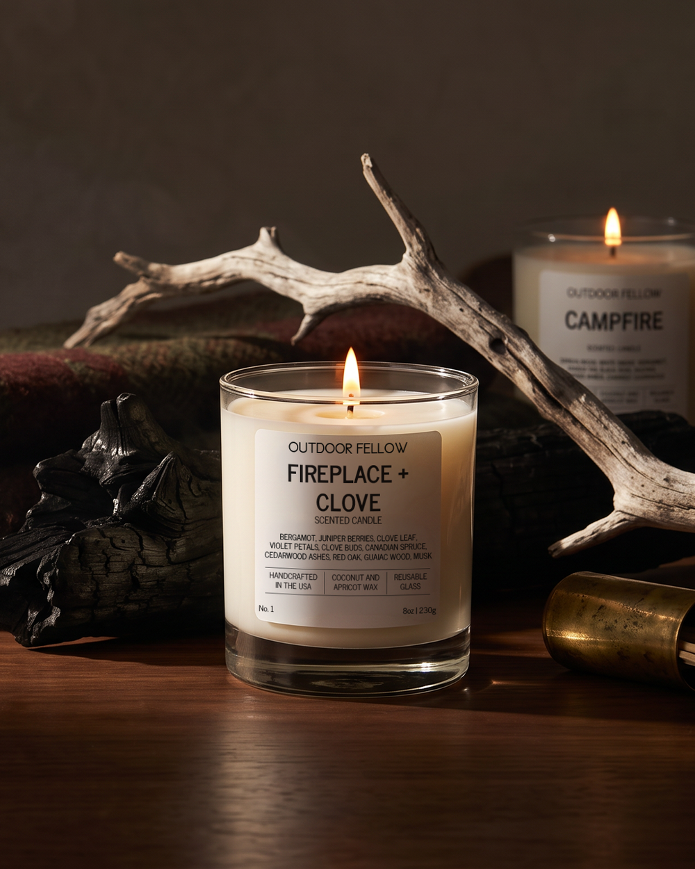 A lit candle in a clear glass jar labeled 'OUTDOOR FELLOW FIREPLACE + CLOVE' sits on a wooden surface, styled with antlers, dark wood pieces, and another candle in the background.