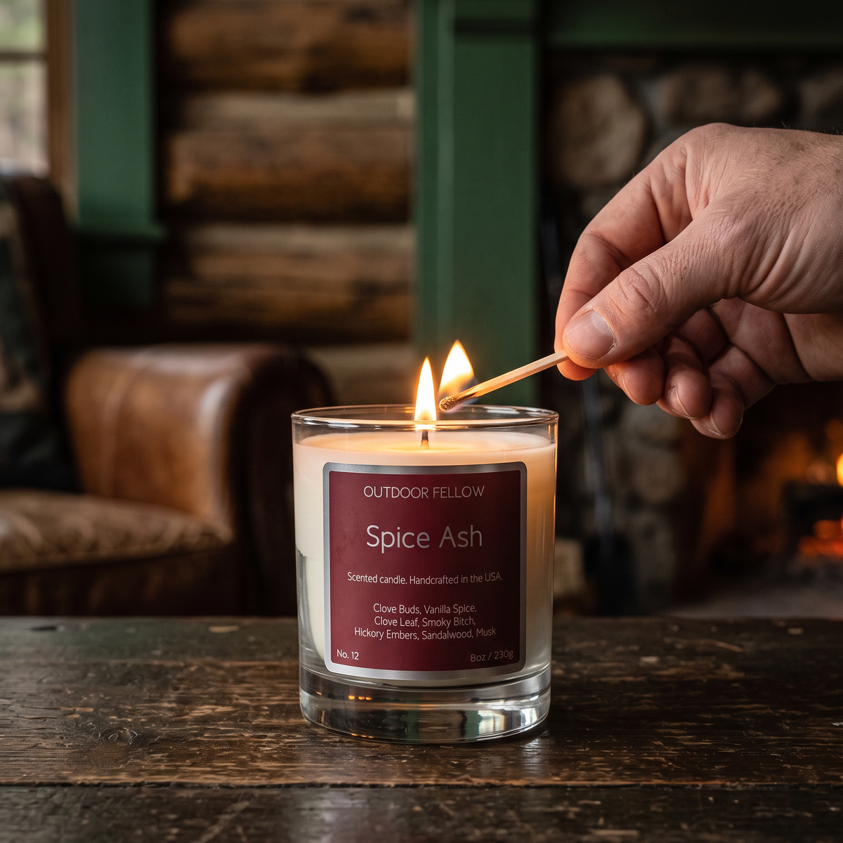 A lit scented candle in a clear glass jar sits on a rustic wooden table, with a person's hand holding a match near the flames. The candle label reads 'OUTDOOR FELLOW Spice Ash'.