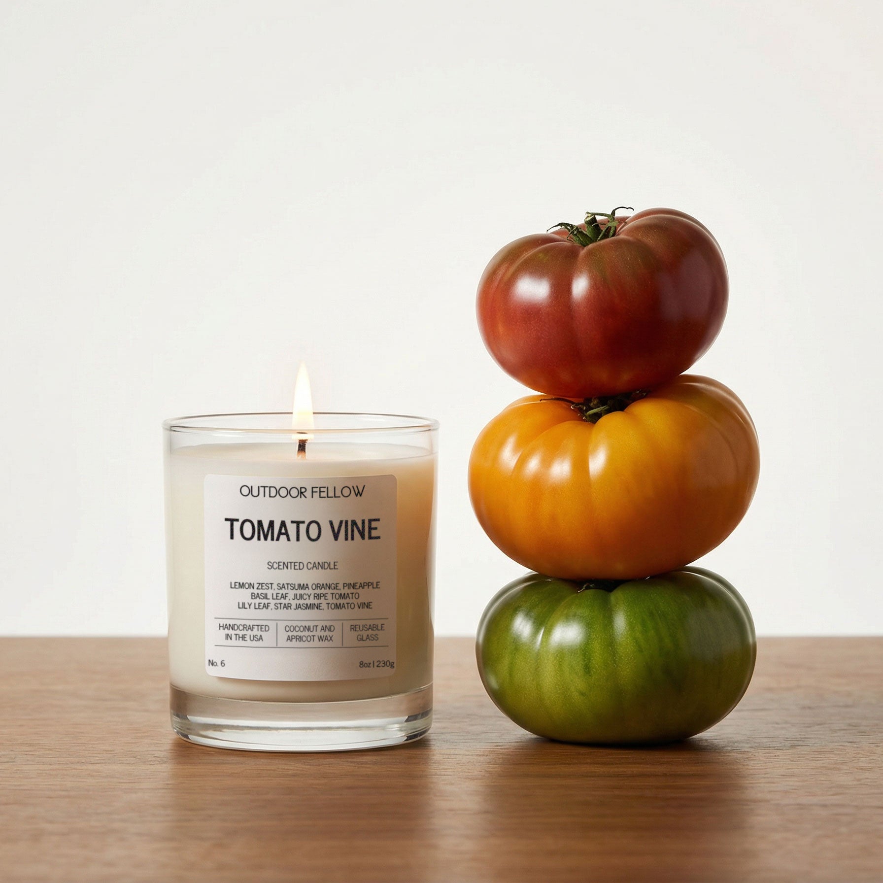 Candle labeled 'Tomato Vine' next to three stacked tomatoes on a wooden surface.