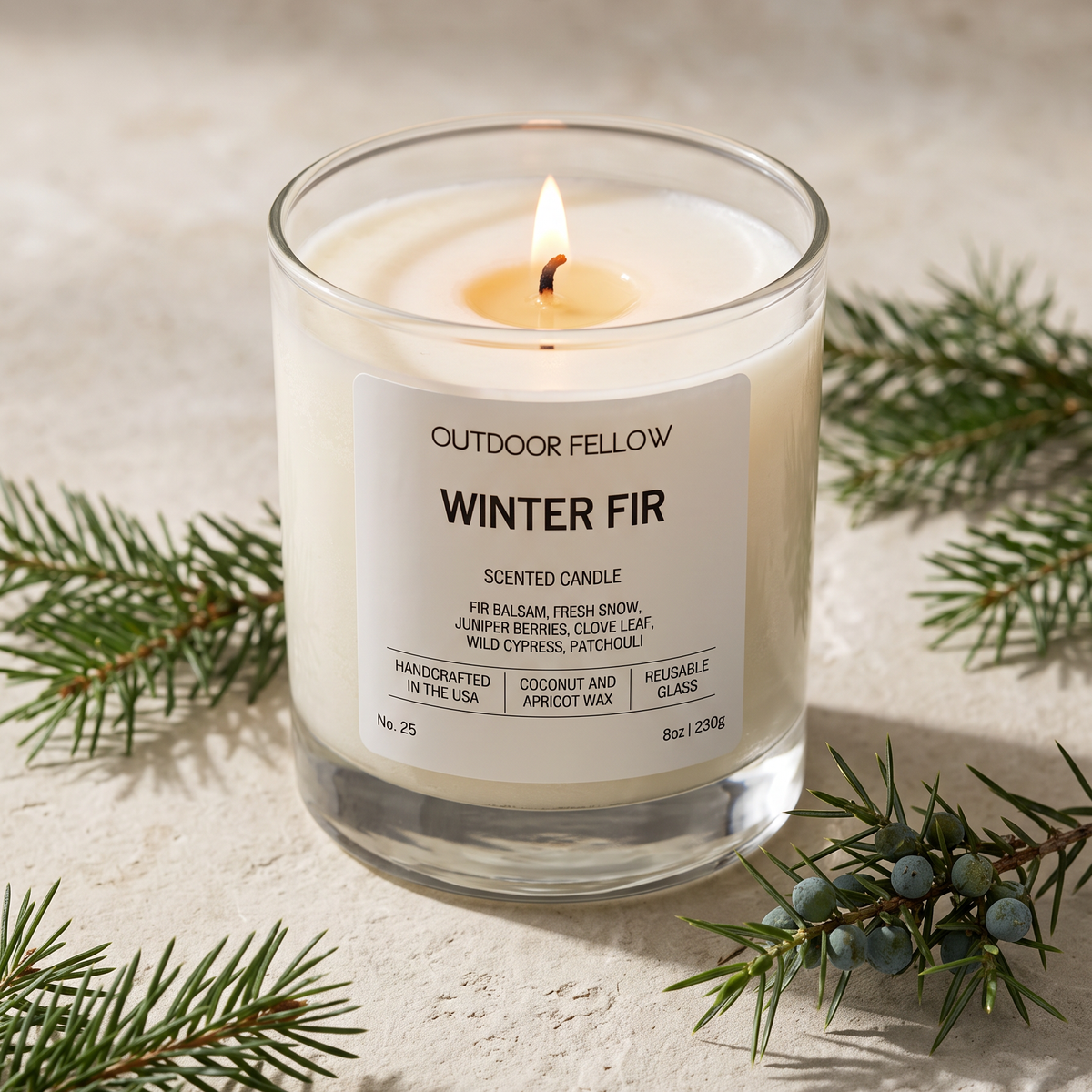 A lit scented candle in a clear glass jar with a white label reading 'OUTDOOR FELLOW WINTER FIR,' styled on a light stone surface with pine branches around it.