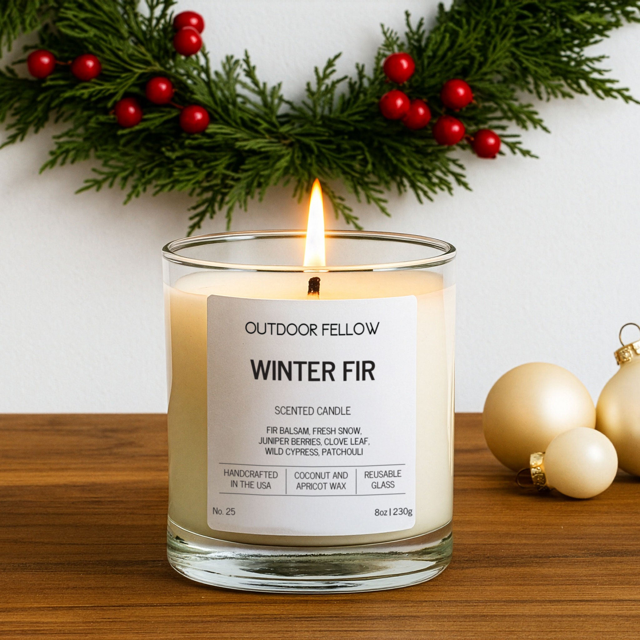Candle labeled 'Outdoor Fellow Winter Fir' with decorative greenery and ornaments on a wooden surface.