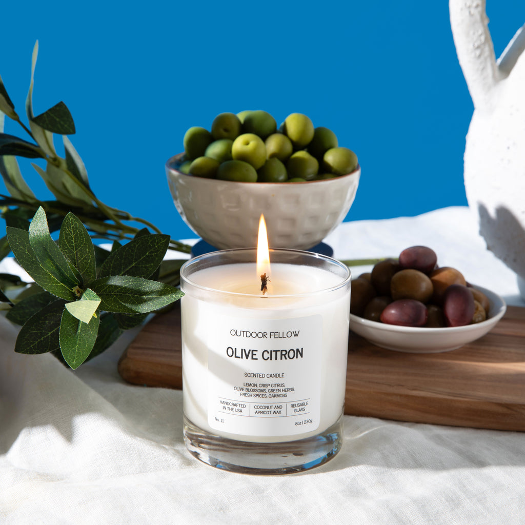  A lit Outdoor Fellow Olive Citron scented candle sits on a white linen cloth surrounded by Mediterranean-inspired elements. To the left, a bowl of lemons and sprigs of olive branches with dark olives rest on a wooden surface. Behind the candle are ceramic bowls filled with green and multicolored olives, and to the right is a white, textured ceramic pitcher. The backdrop is a vibrant blue, creating a bright and fresh atmosphere.