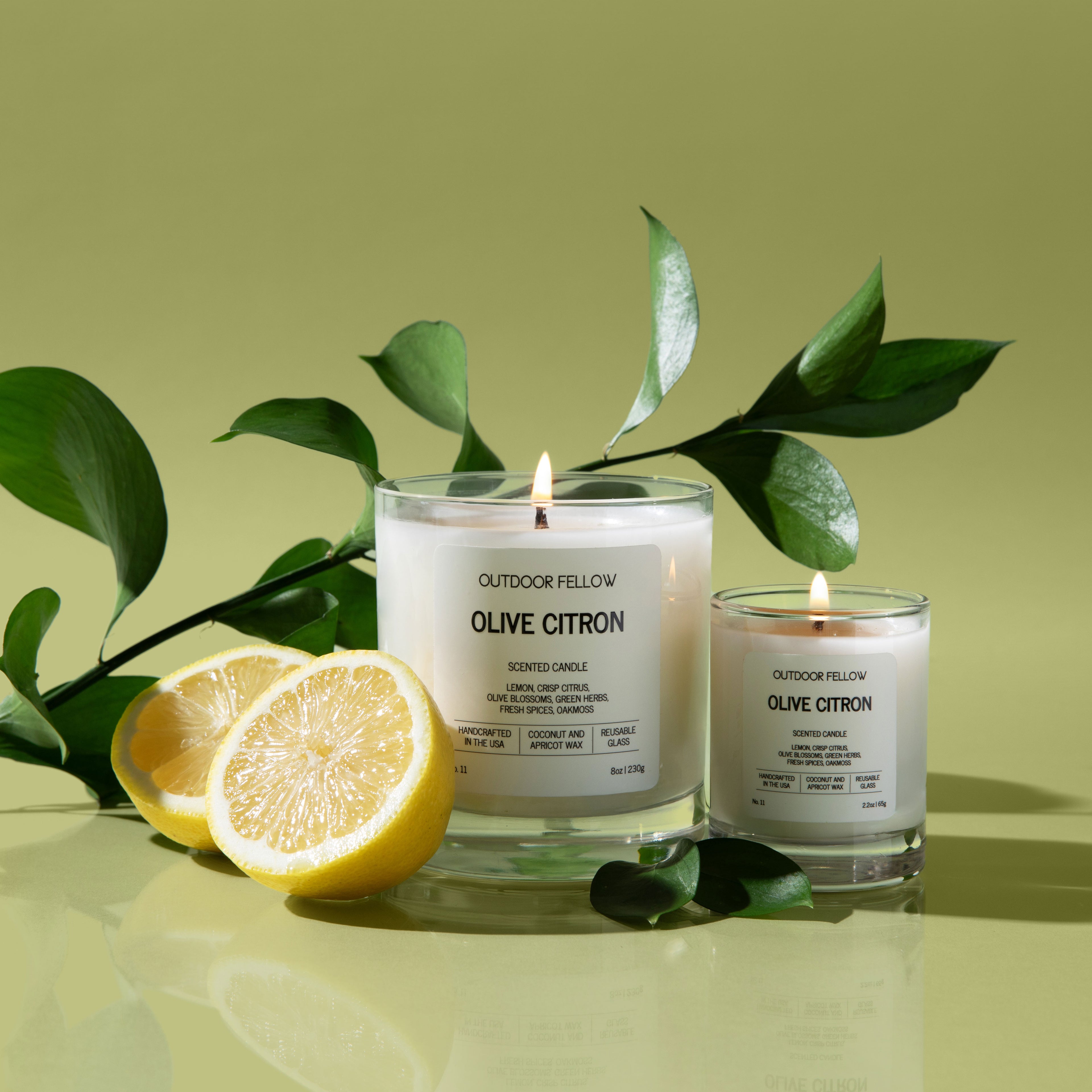 Two lit white candles labeled “Outdoor Fellow Olive Citron” sit on a glossy surface with lemon halves and green leaves nearby. The background is a soft olive green, creating a fresh and minimal look.