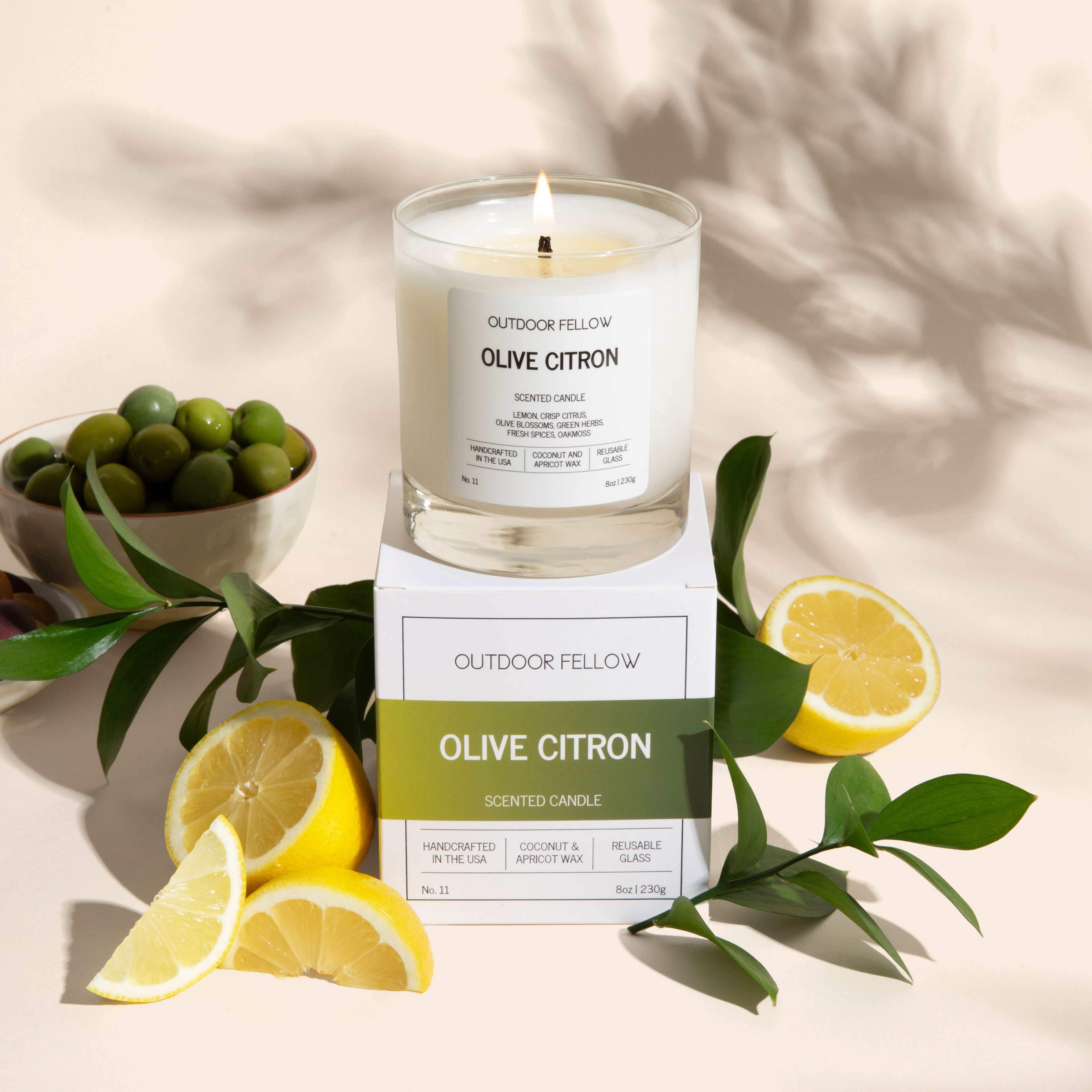 A lit candle labeled “Outdoor Fellow Olive Citron” sits on top of its product box. Around it are lemon slices, green leaves, and bowls of green and brown olives. The background is a soft beige with leafy shadows.