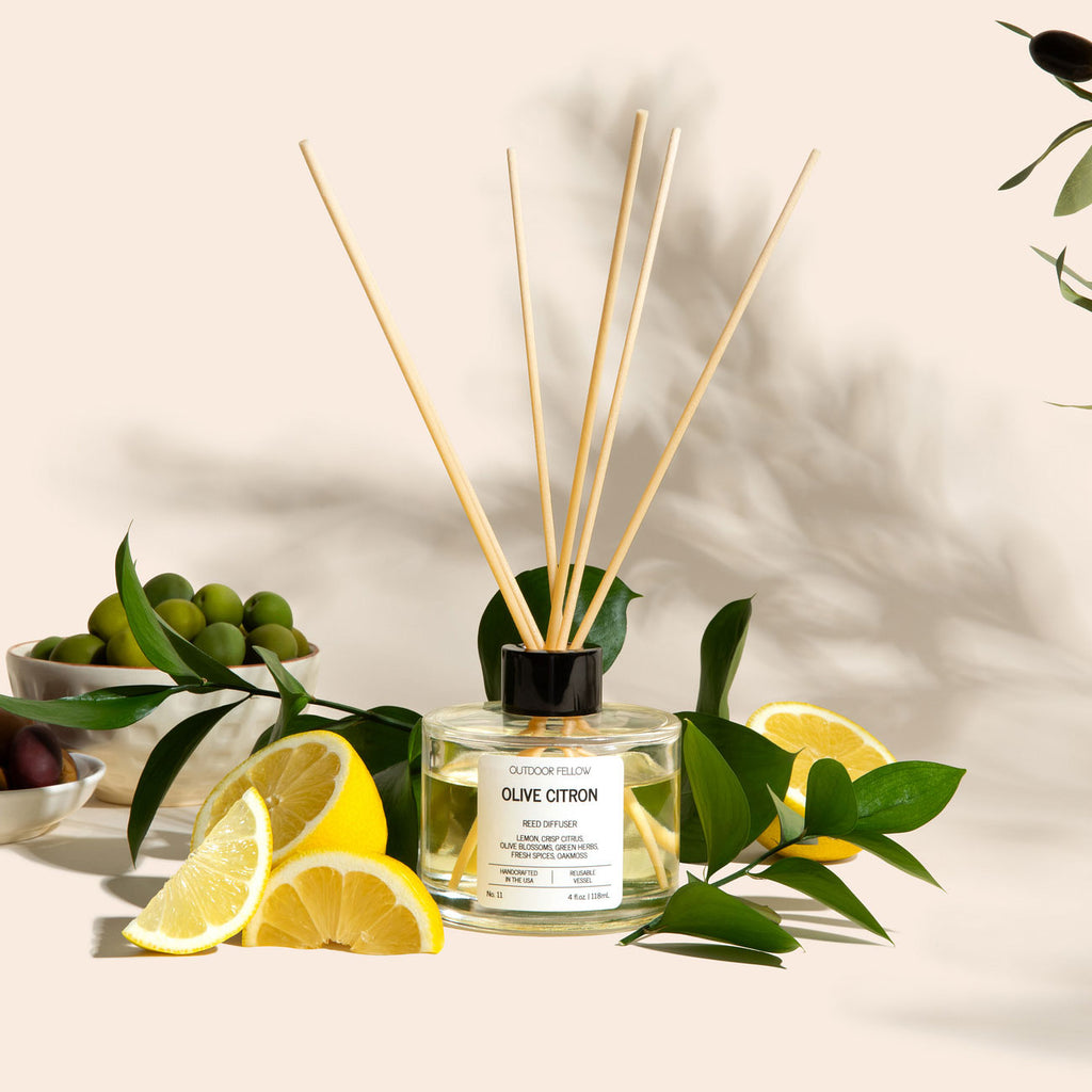 Outdoor Fellow Olive Citron reed diffuser surrounded by sliced lemons, green leaves, and bowls of olives on a neutral background with soft shadows.