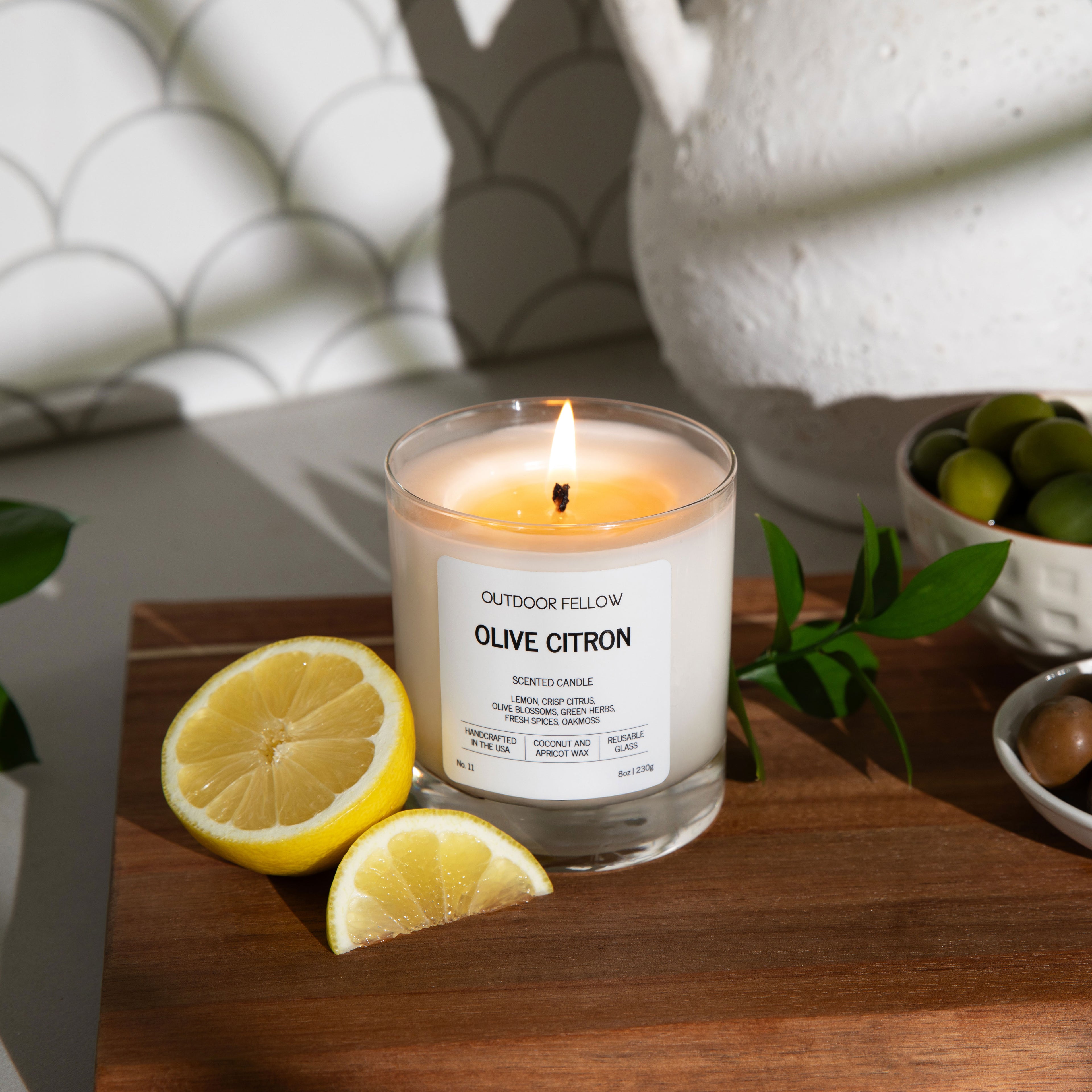 A lit “Outdoor Fellow Olive Citron” candle sits on a wooden board next to sliced lemons, green leaves, and small bowls of green and brown olives. The background features a white textured pitcher and a tiled kitchen wall with scalloped patterns.