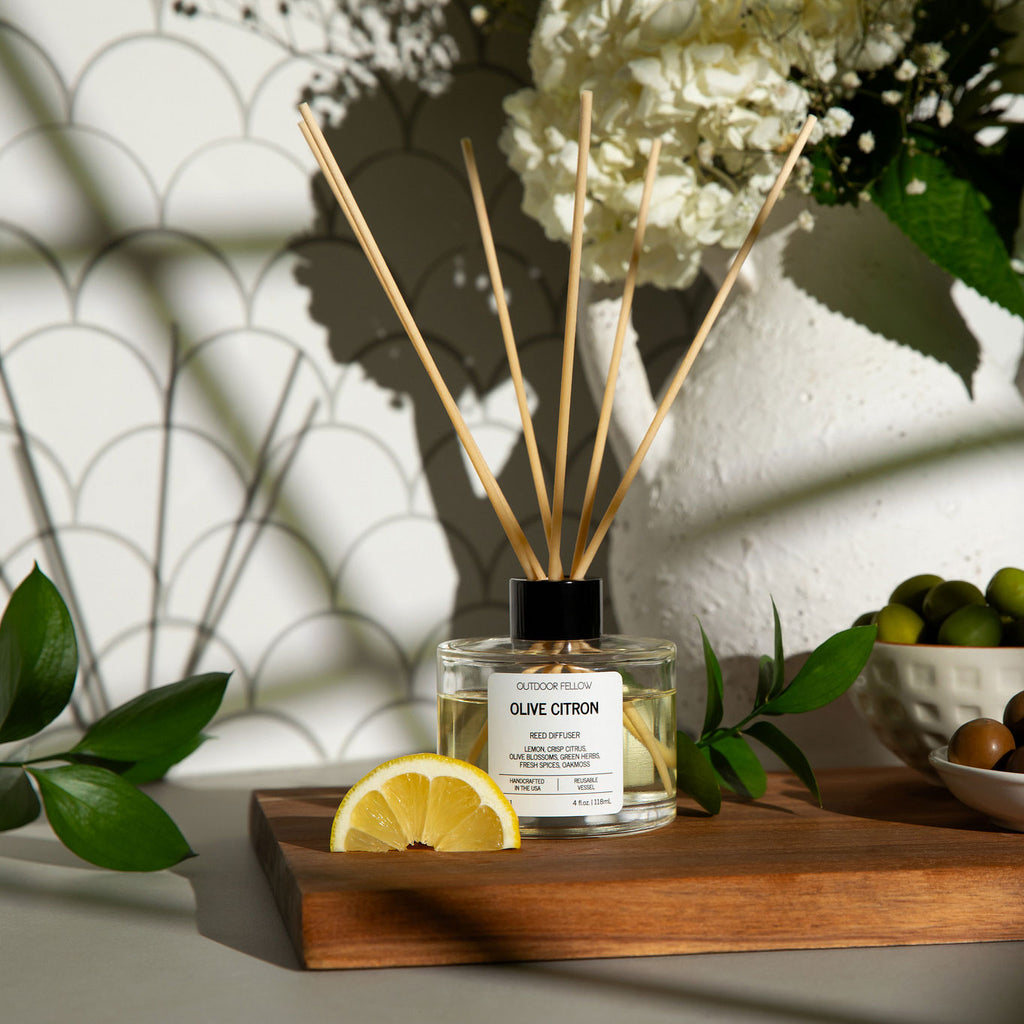 Outdoor Fellow Olive Citron reed diffuser on a wooden board with lemon slices and olives, surrounded by greenery and white flowers in natural sunlight.