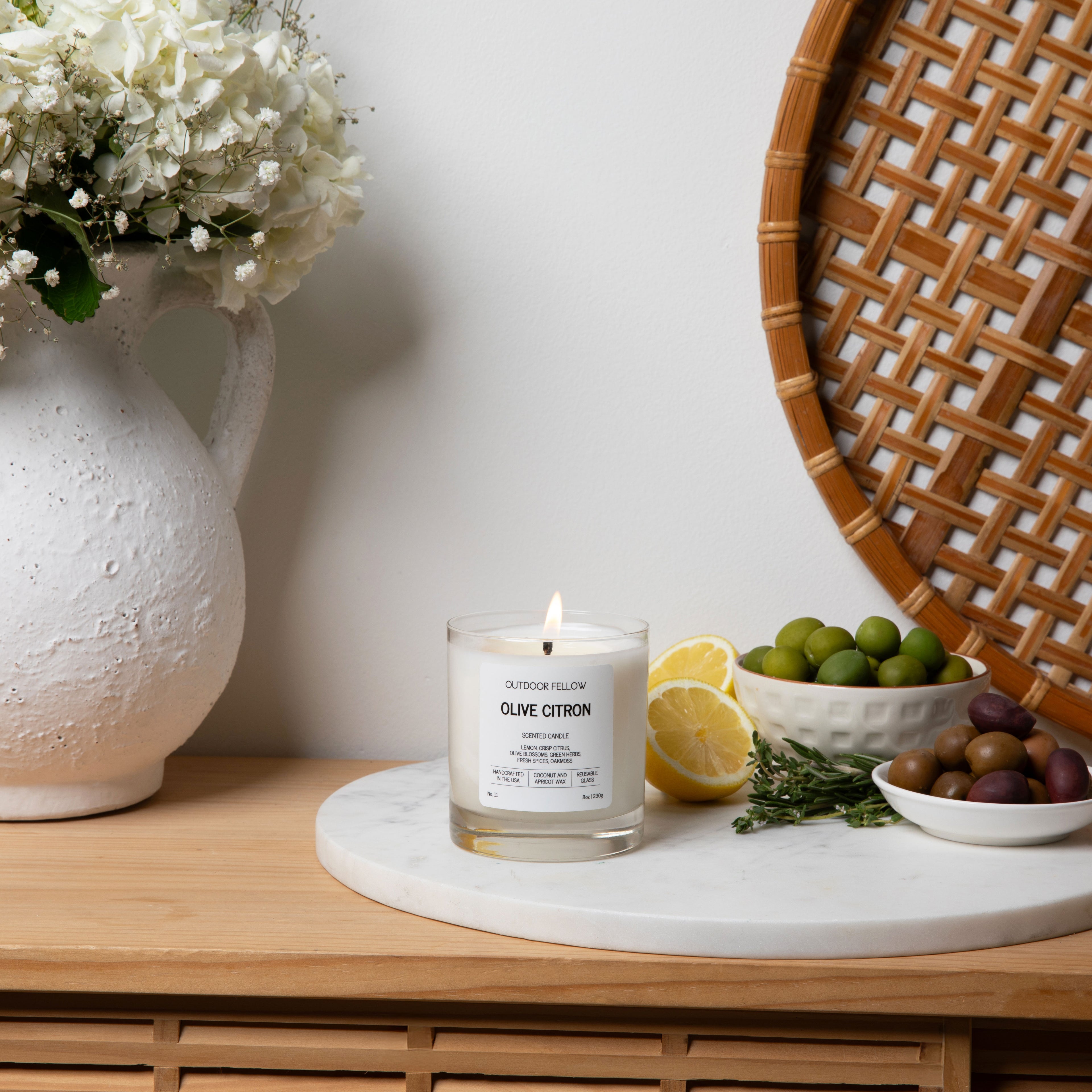 A lit “Outdoor Fellow Olive Citron” candle sits on a round white marble tray, surrounded by lemon slices, fresh herbs, and small bowls of green and brown olives. To the left is a large white textured vase filled with white flowers, and to the right is a woven rattan wall piece against a white background.