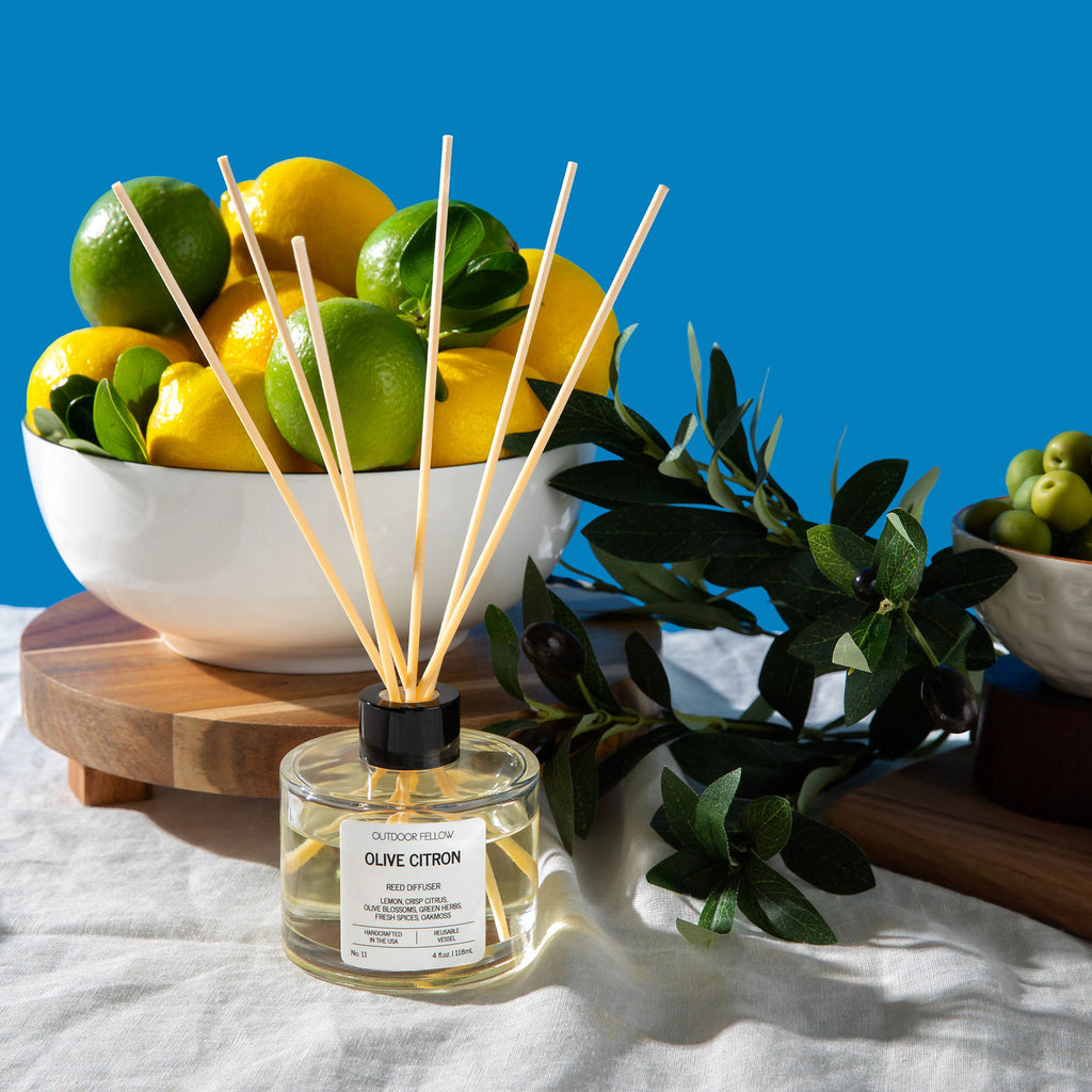 Outdoor Fellow Olive Citron reed diffuser displayed in front of a bowl of lemons and limes, with olive branches and green olives surrounding it on a sunlit table.