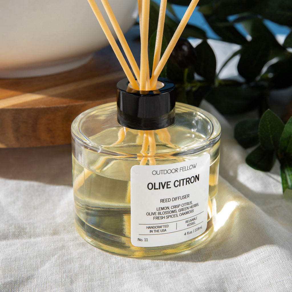 Close-up of the Outdoor Fellow Olive Citron reed diffuser on a sunlit table, with natural reeds and visible scent label.