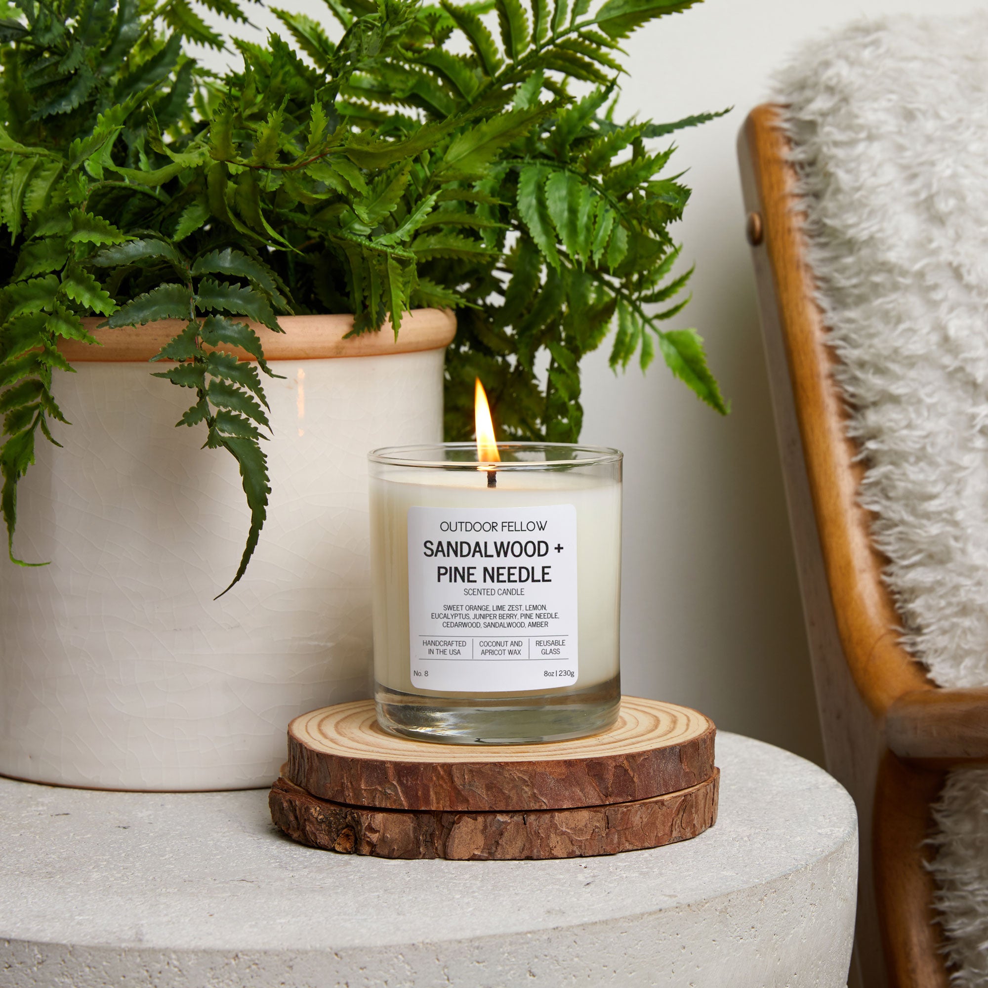 Candle labeled 'Sandalwood + Pine Needle' on a wooden coaster with a plant in the background.