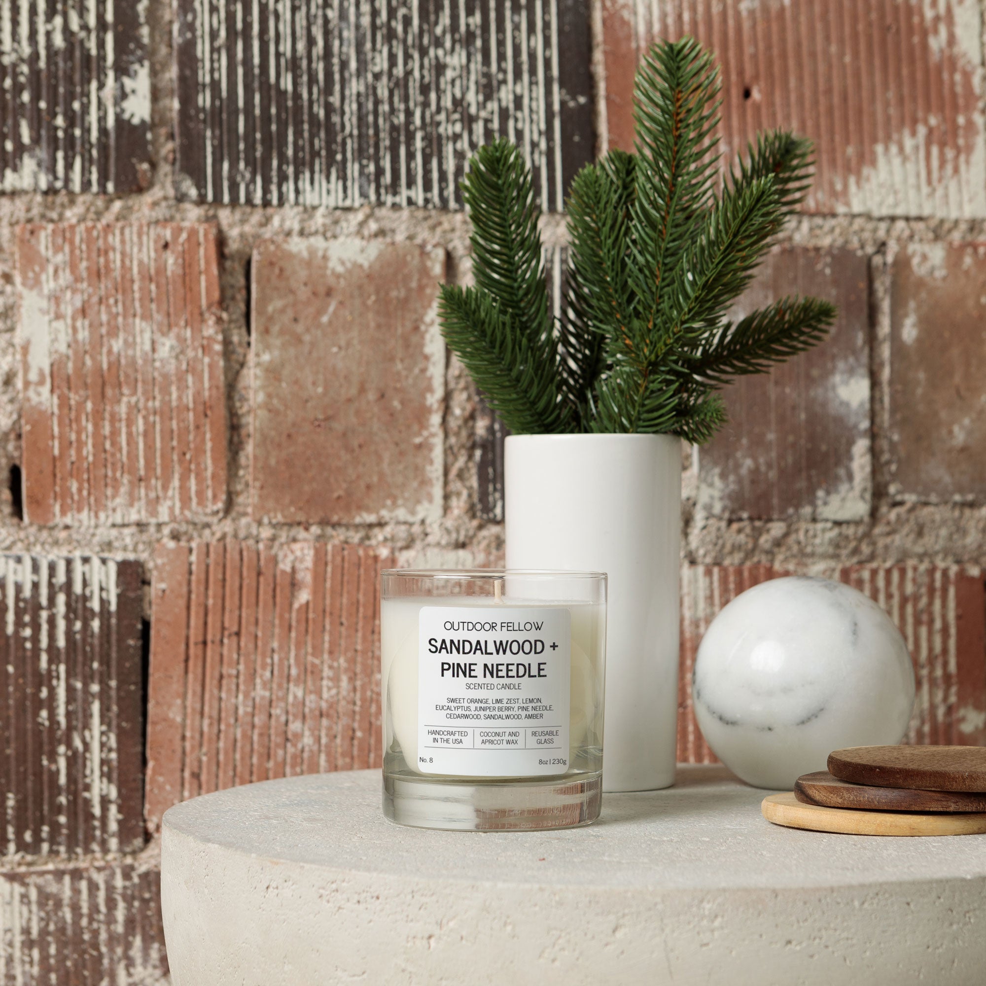 Candle labeled 'Sandalwood + Pine Needle' on a surface with a plant and decorative items against a brick wall.