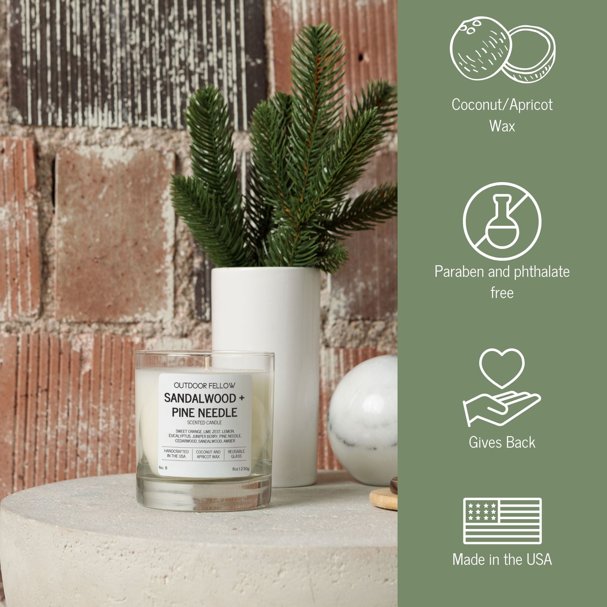 An Outdoor Fellow Sandalwood + Pine Needle candle sits on a stone surface beside a white vase with pine branches and a marble sphere, against a rustic brick wall. On the right, icons with text highlight product details: coconut/apricot wax, paraben and phthalate free, gives back, and made in the USA.