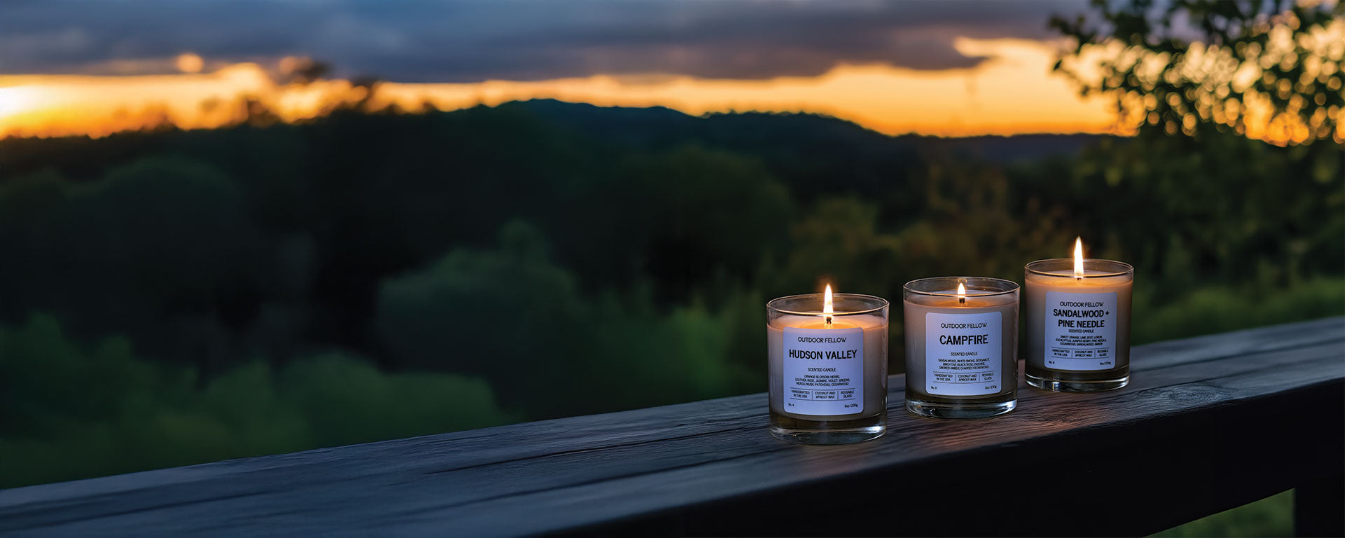 Hudson Valley, Campfire and Sandalwood + Pine Needle candles sitting on a wooden railing with a scenic mountain forest in the background