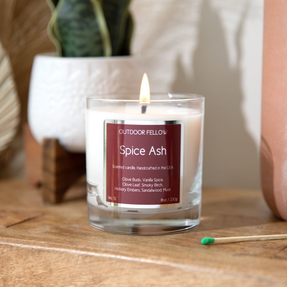 Spice Ash scented candle on a wooden bench with a single match stick in front of it