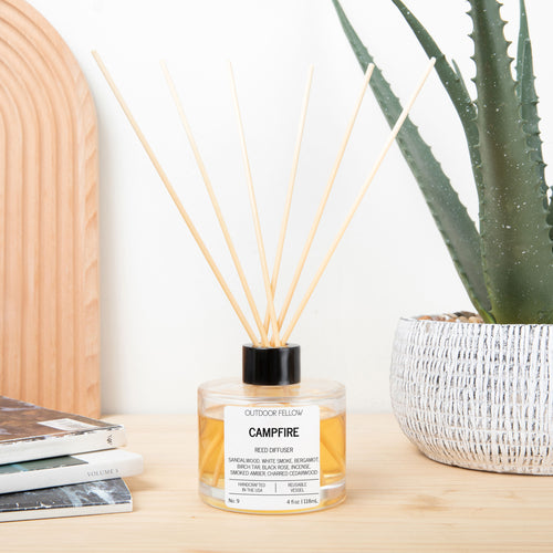 Reed diffuser labeled 'Campfire' on a wooden surface with a plant and books in the background