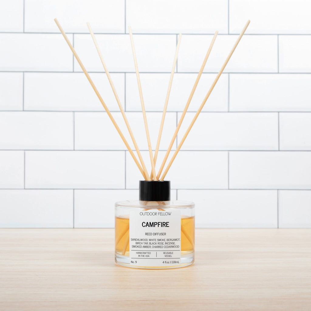 Outdoor Fellow Campfire reed diffuser with amber liquid in a clear glass bottle, black cap, and wooden reeds, placed on a light wood surface in front of white subway tile.
