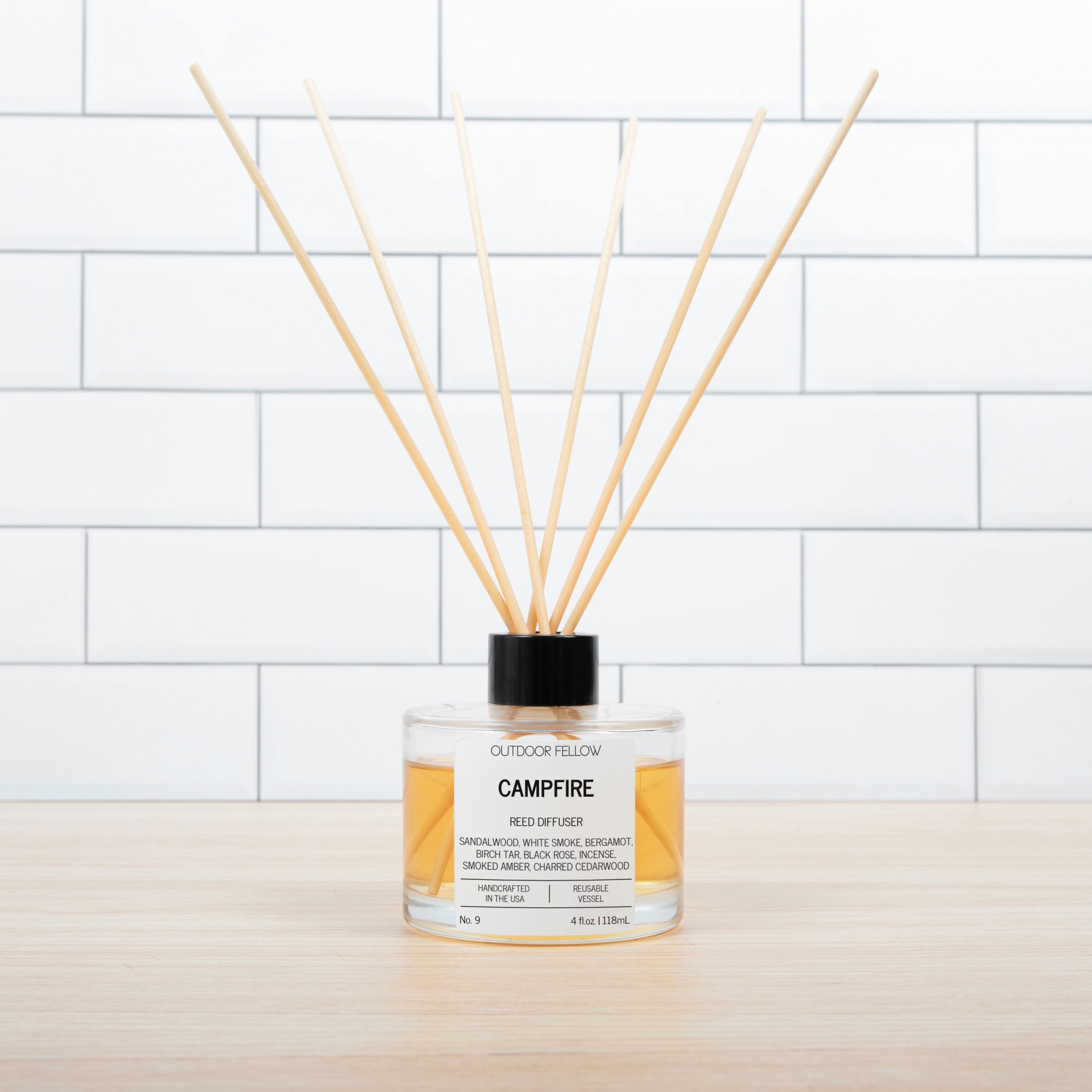 Outdoor Fellow Campfire reed diffuser with amber liquid in a clear glass bottle, black cap, and wooden reeds, placed on a light wood surface in front of white subway tile.