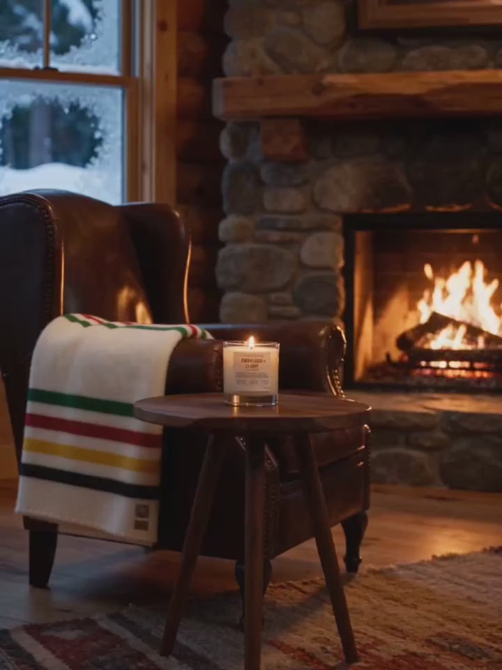 Cozy cabin scene with a lit Outdoor Fellow candle on a wooden side table beside a leather armchair draped with a striped wool blanket, with a glowing stone fireplace and snowy view in the background.
