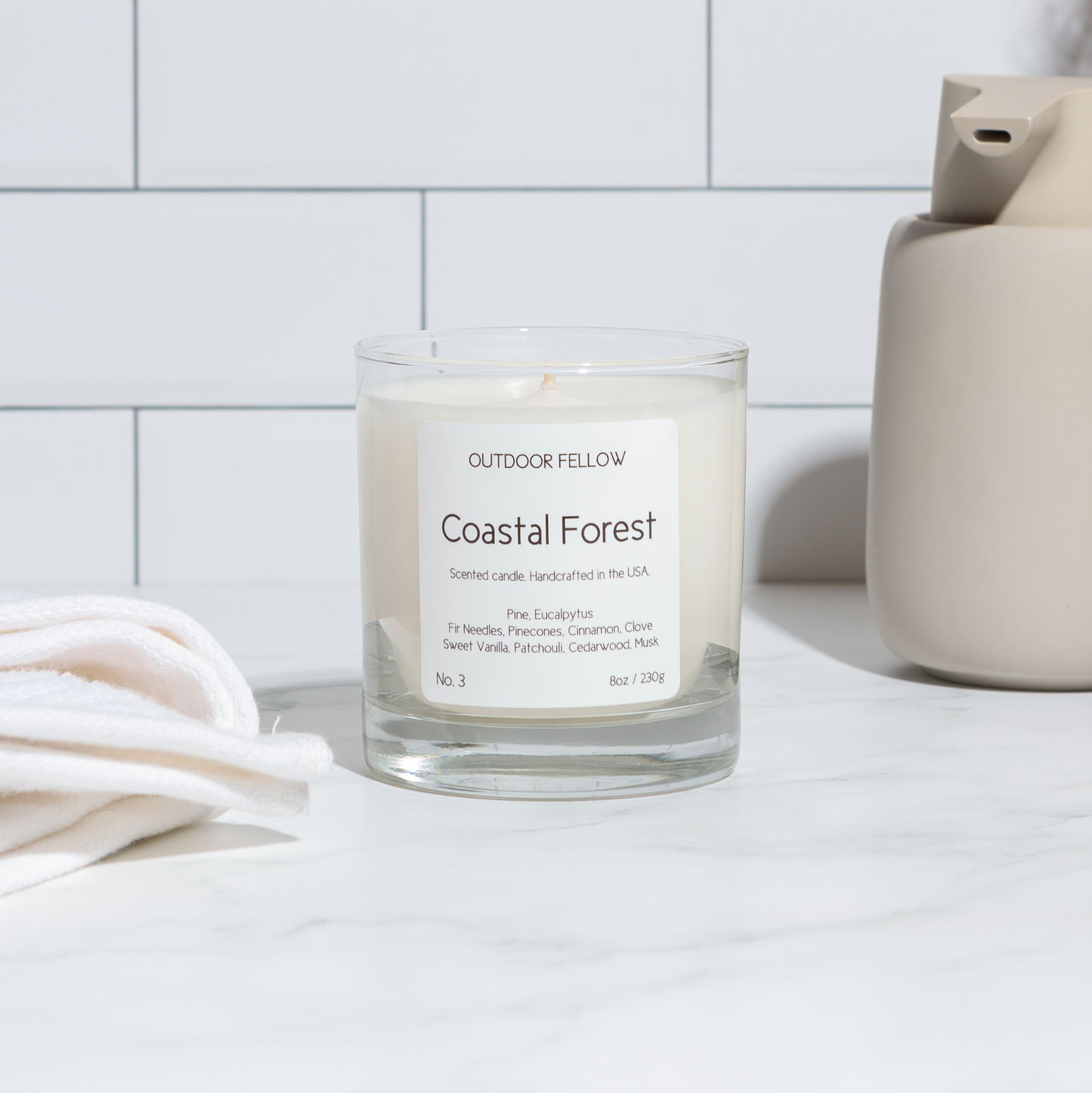 Coastal Forest scented candle next to a towel and soap dispenser in a bathroom setting