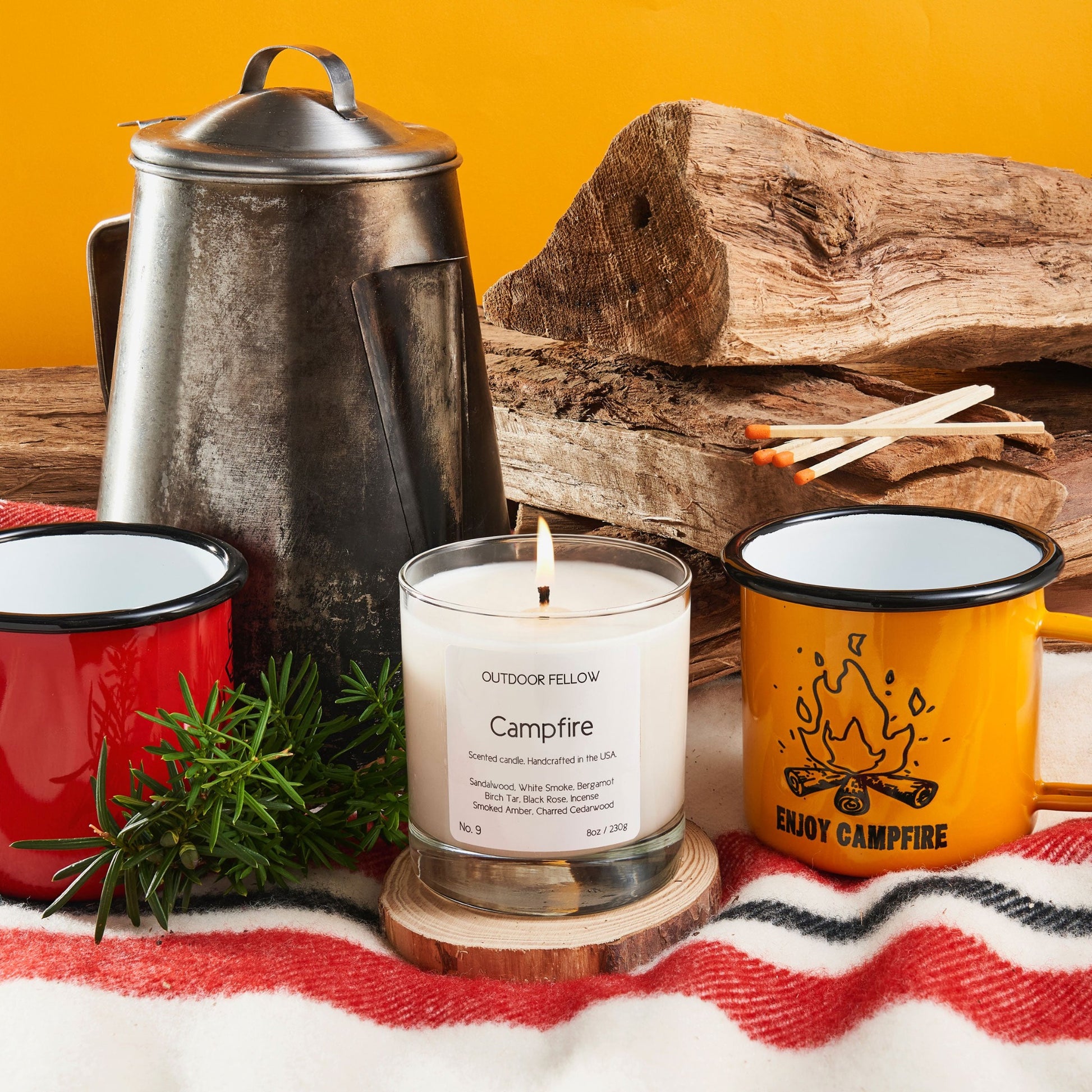 Campfire scented candle next to coffee mugs, stacked wood and a rustic coffee pot