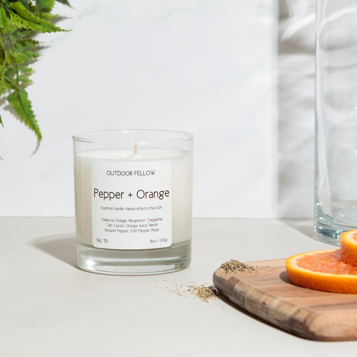 Pepper and Orange scented candle next to orange slices and ground pepper on cutting board