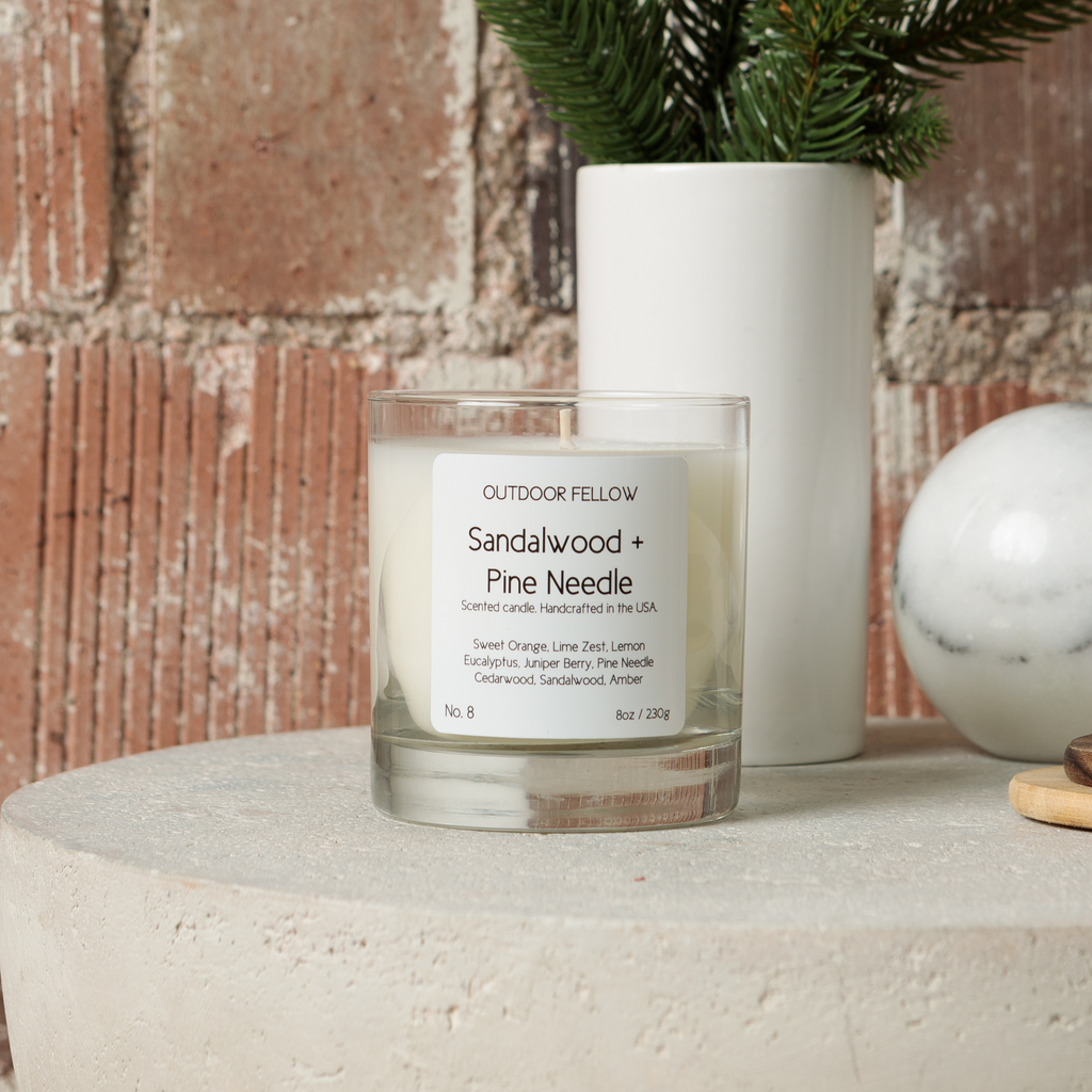 Sandalwood and Pine Needle scented candle on concrete table with brick background next to a vase with pine clippings